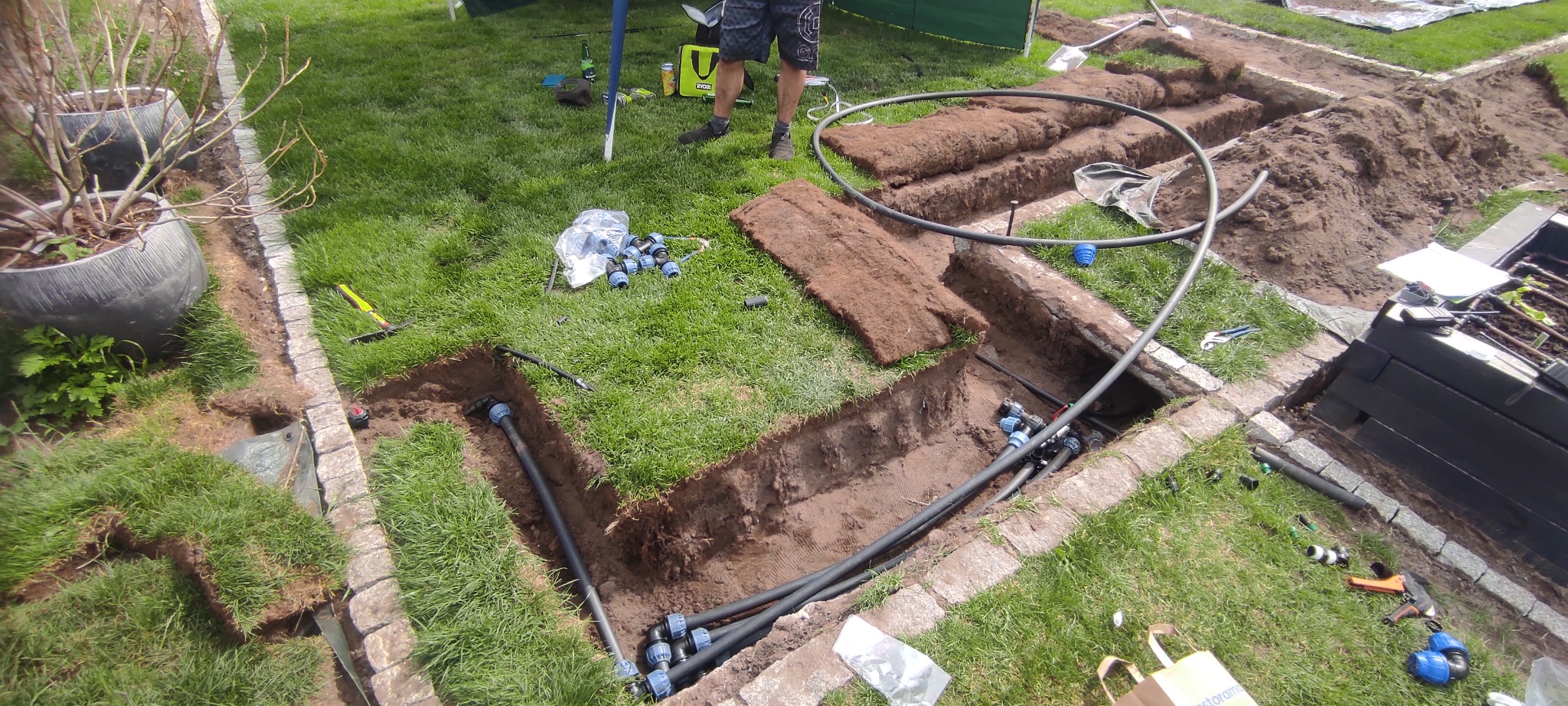 Wykop w trawniku z ułożonymi czarnymi rurami i złączkami systemu nawadniania, obok leżą fragmenty darni, widoczne narzędzia i elementy instalacji