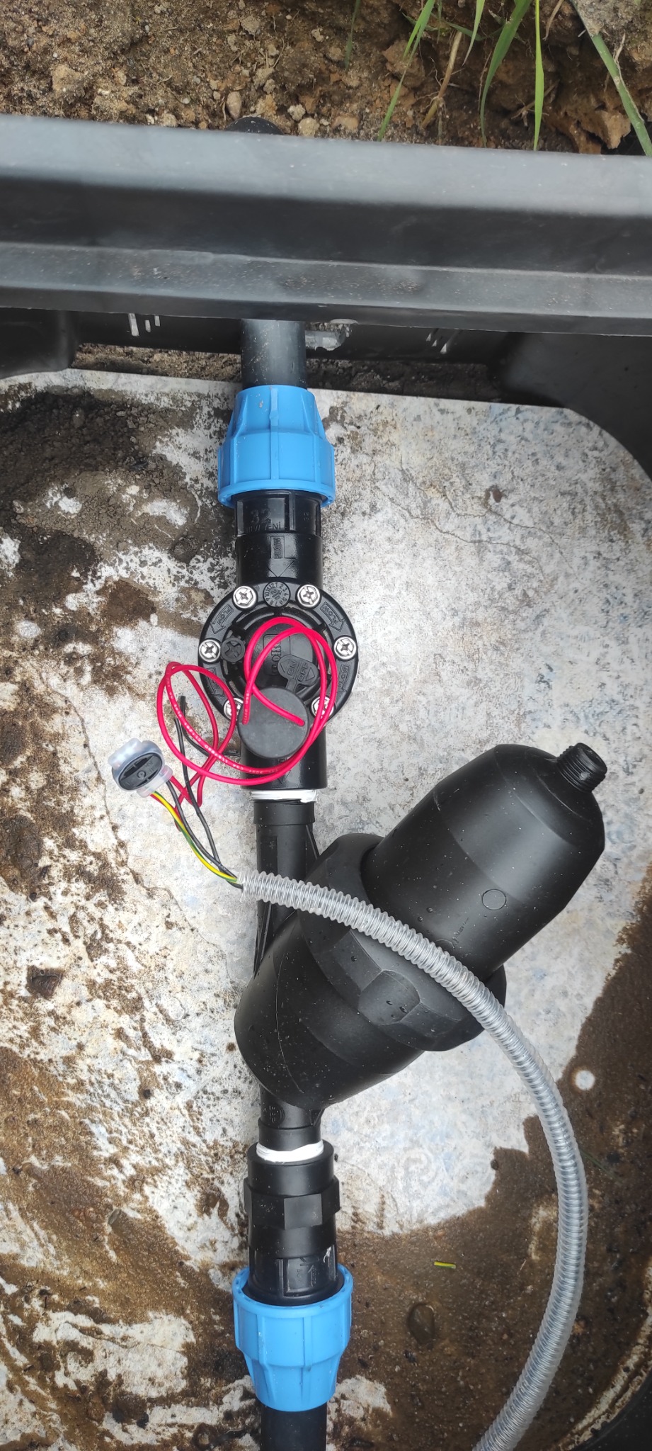 Elementy systemu nawadniania, z widocznymi złączkami, przewodami i filtrem, umieszczone w plastikowej skrzynce w ziemi.