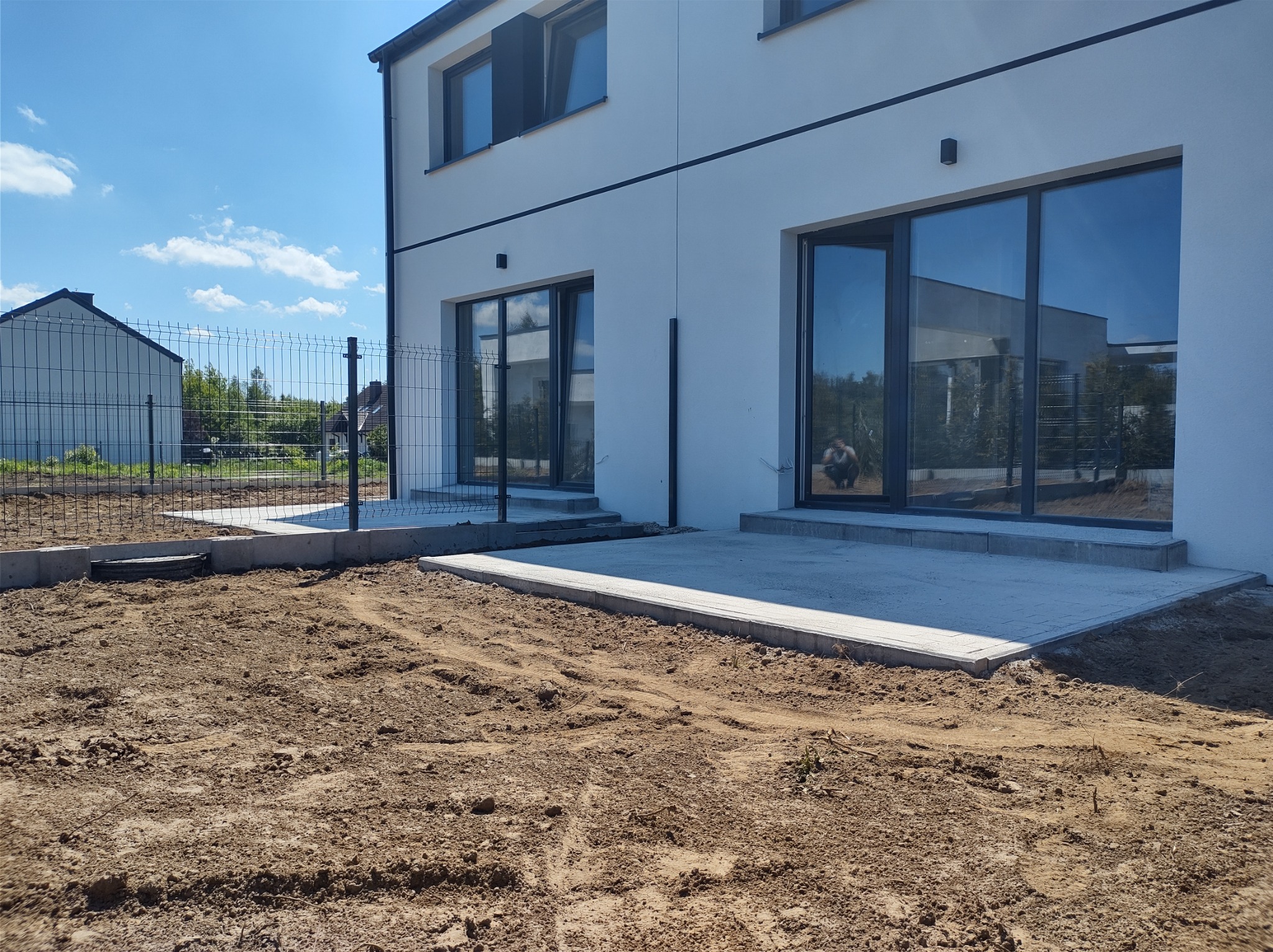 Nowoczesny dom z widocznym fragmentem nowego tarasu z jasnej kostki brukowej i ogrodzeniem panelowym, teren przygotowany pod dalsze prace.