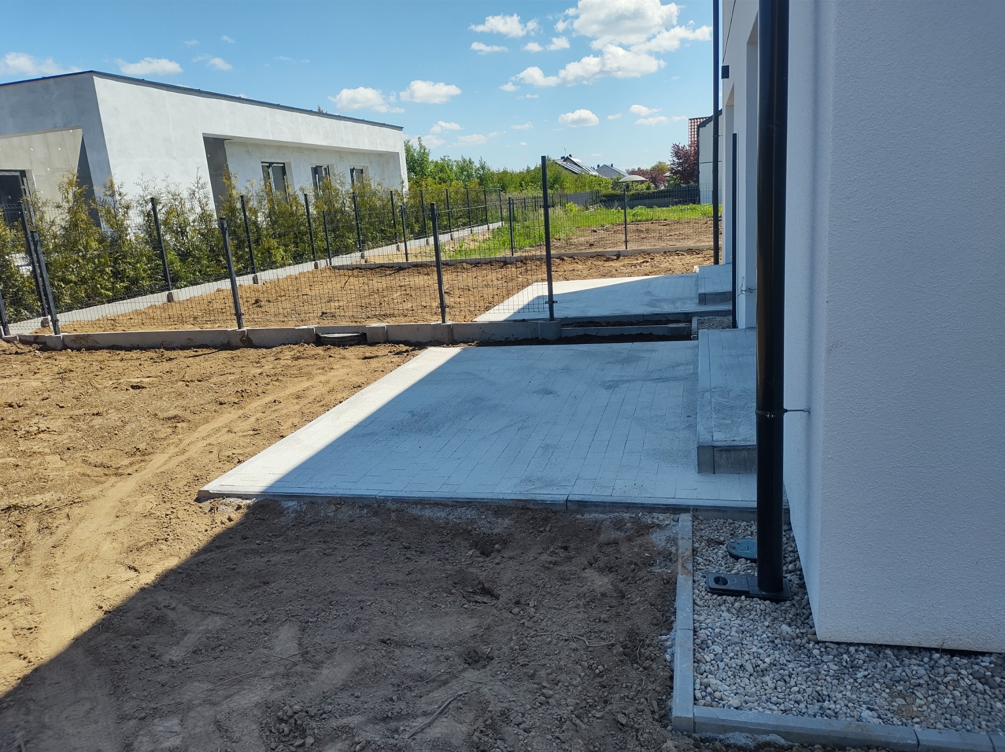 Świeżo ułożona, jasnoszara kostka brukowa tworząca ścieżkę wokół domu w Białogardzie, z widocznym przygotowanym terenem pod dalsze prace i fragmentem ogrodzenia.