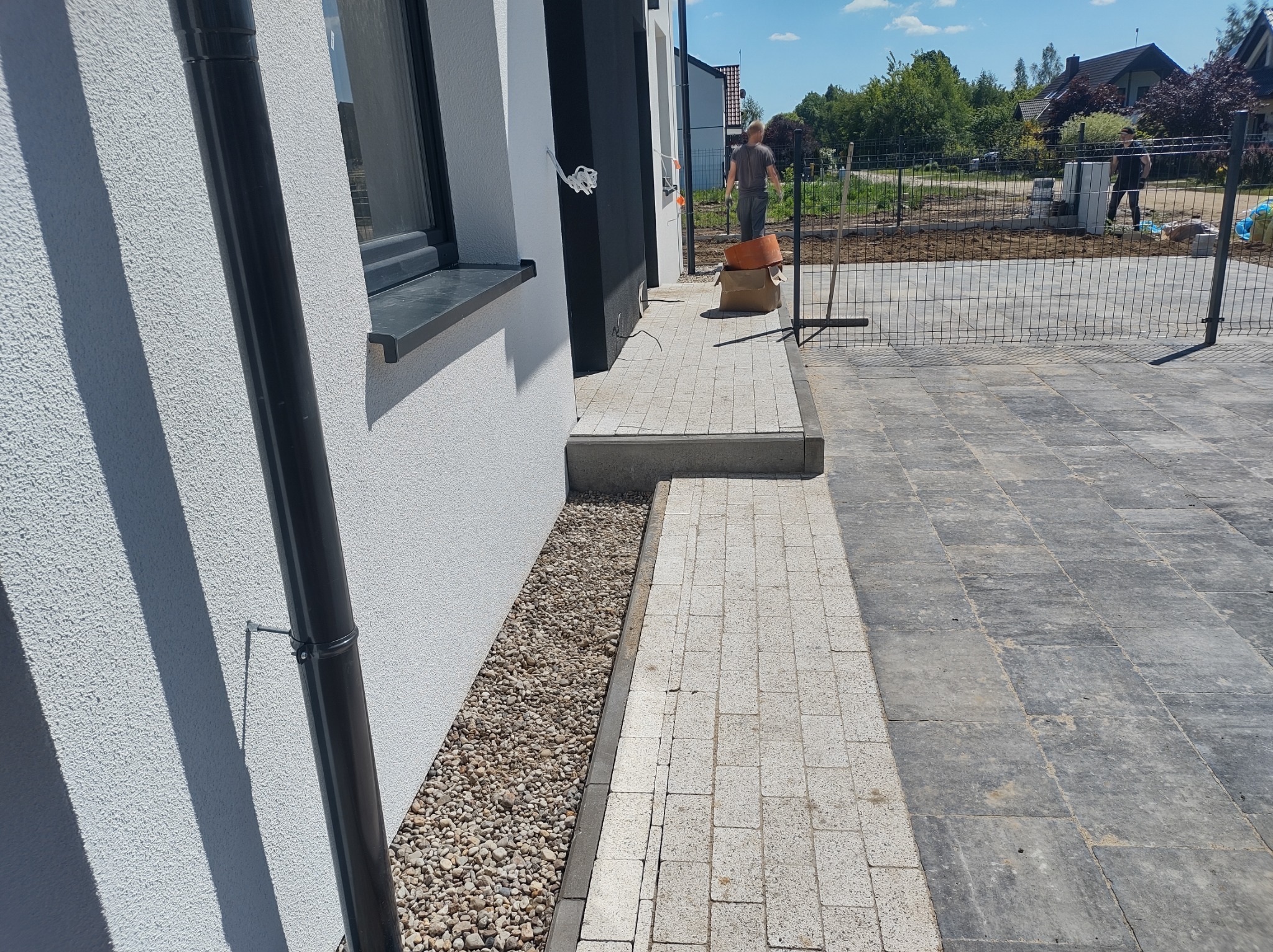 Świeżo ułożona kostka brukowa przed domem z widocznym fragmentem montowanego ogrodzenia w tle.