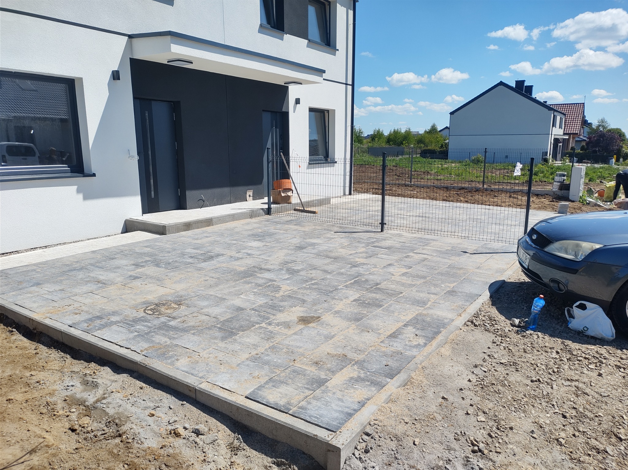 Ułożona szara kostka brukowa przed nowoczesnym domem z czarnymi elementami, widoczny fragment ogrodzenia z siatki i samochód osobowy z butelką wody obok.
