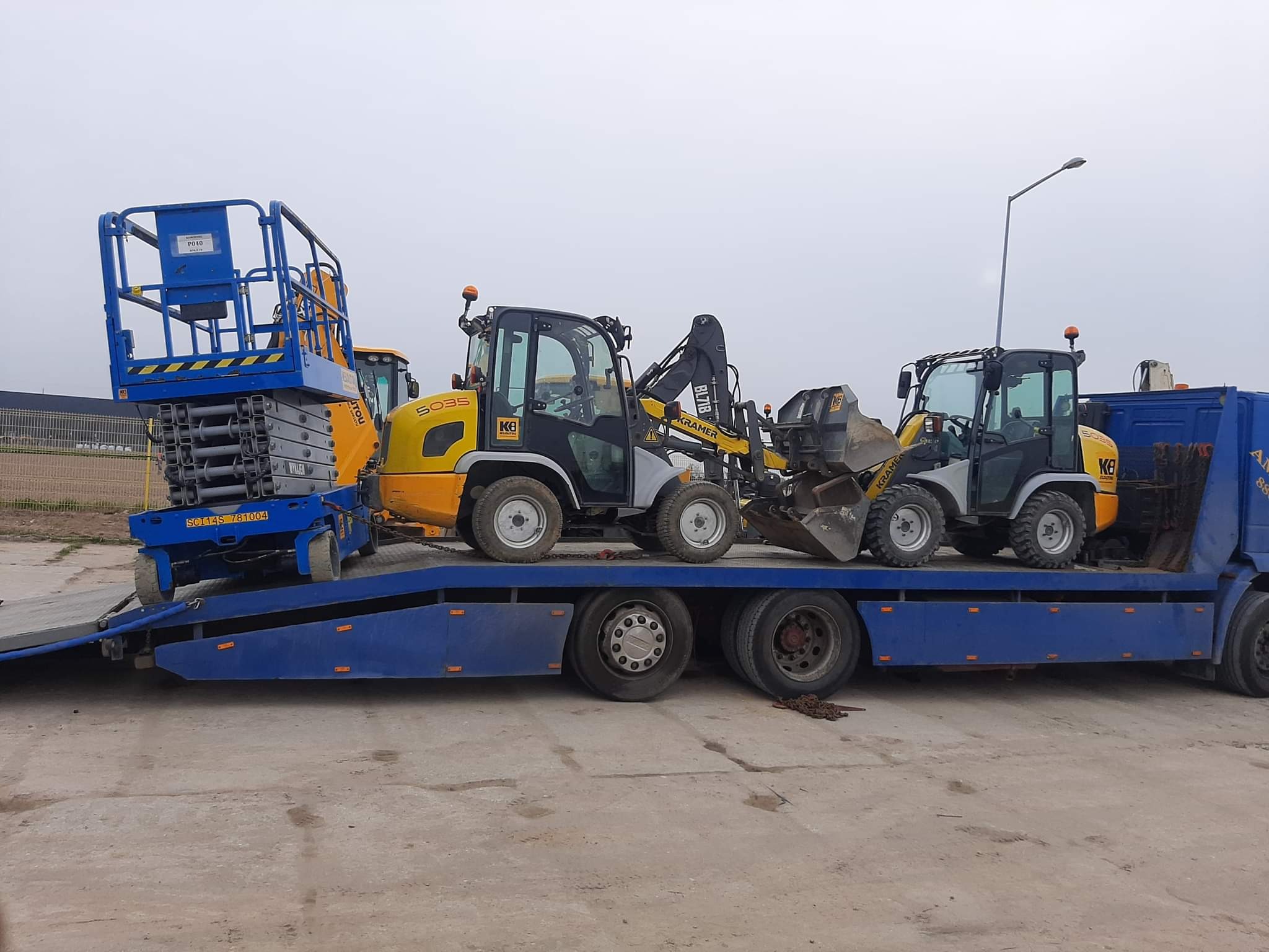 Niebieska laweta z rozłożoną rampą przewozi podnośnik koszowy i dwie mini ładowarki Kramer. Widoczne łańcuchy zabezpieczające ładunek.