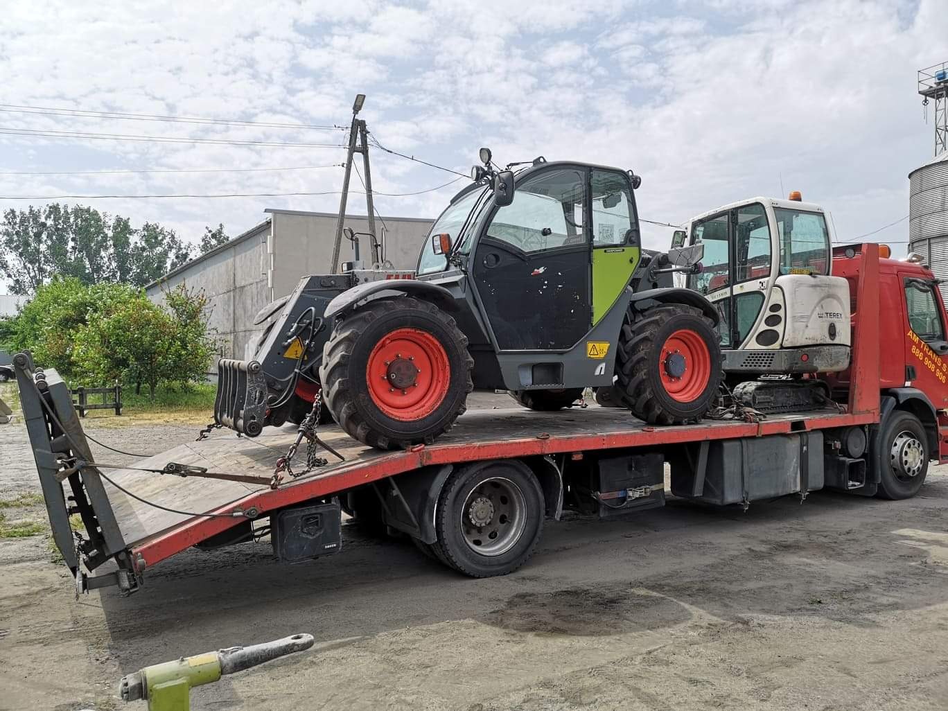 Czerwona laweta przewozi szaro-zielonego teleskopowego ładowacza Terex i białą mini-koparkę na tle silosów zbożowych.