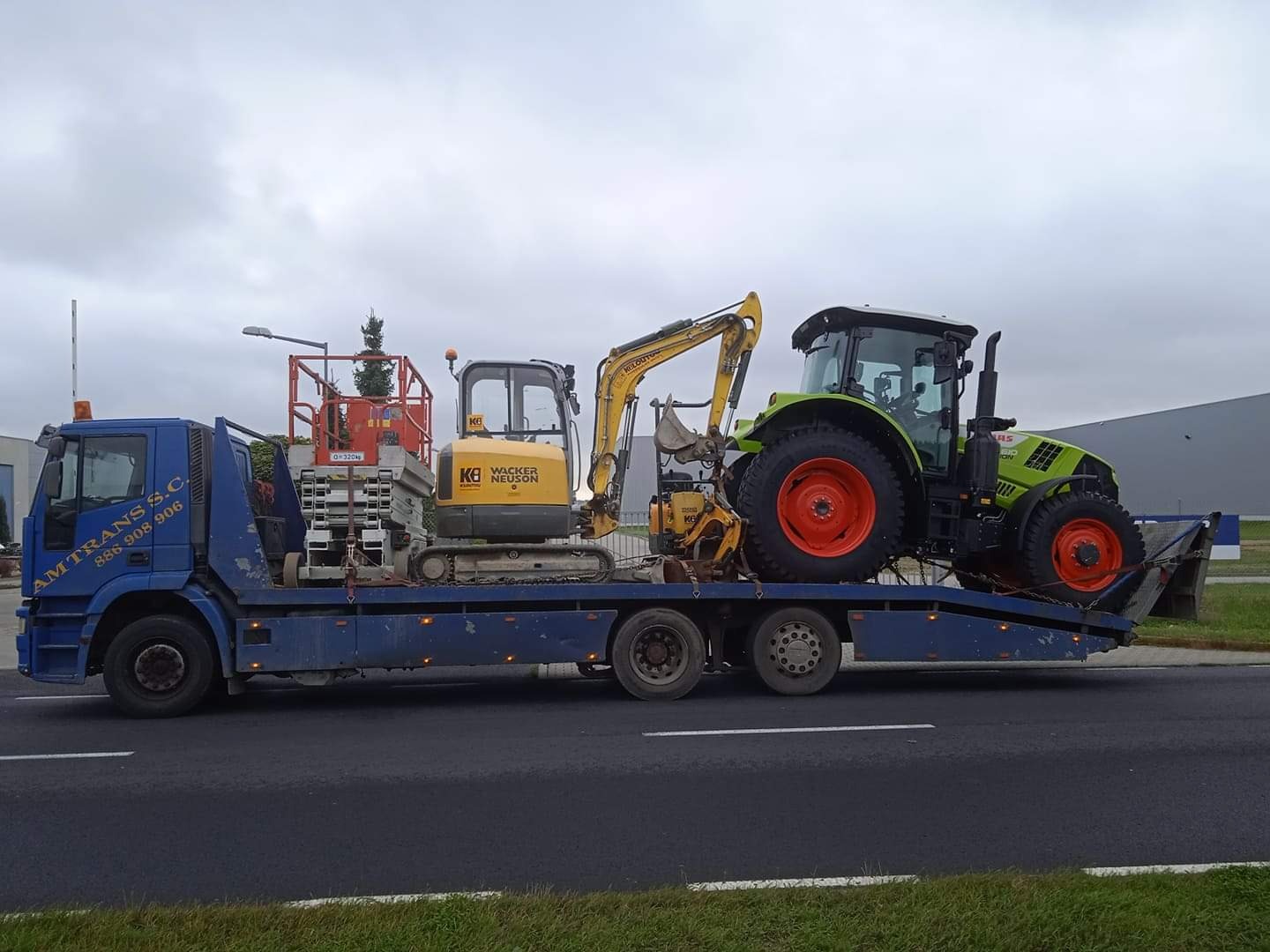 Niebieska ciężarówka z logo 'AM TRANS S.C.' przewozi na lawecie żółtą mini koparkę Wacker Neuson, podnośnik koszowy i zielony traktor Claas z pomarańczowymi felgami na tle pochmurnego nieba...