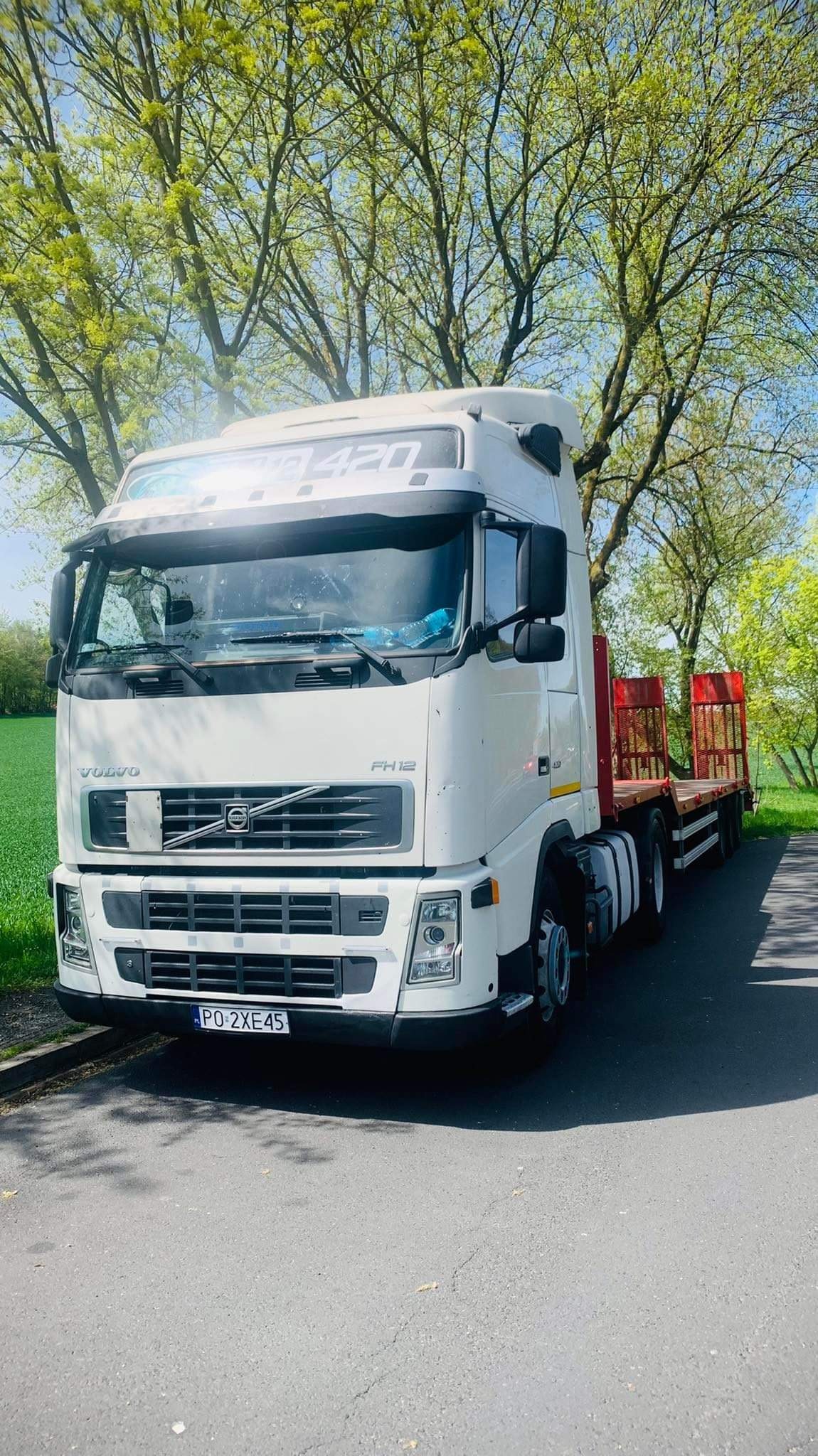 Biała ciężarówka Volvo FH12 z niskopodwoziową platformą, zaparkowana na asfalcie w słoneczny dzień, z widocznymi drzewami w tle. Tablica rejestracyjna z przodu pojazdu to PO 2XE45.