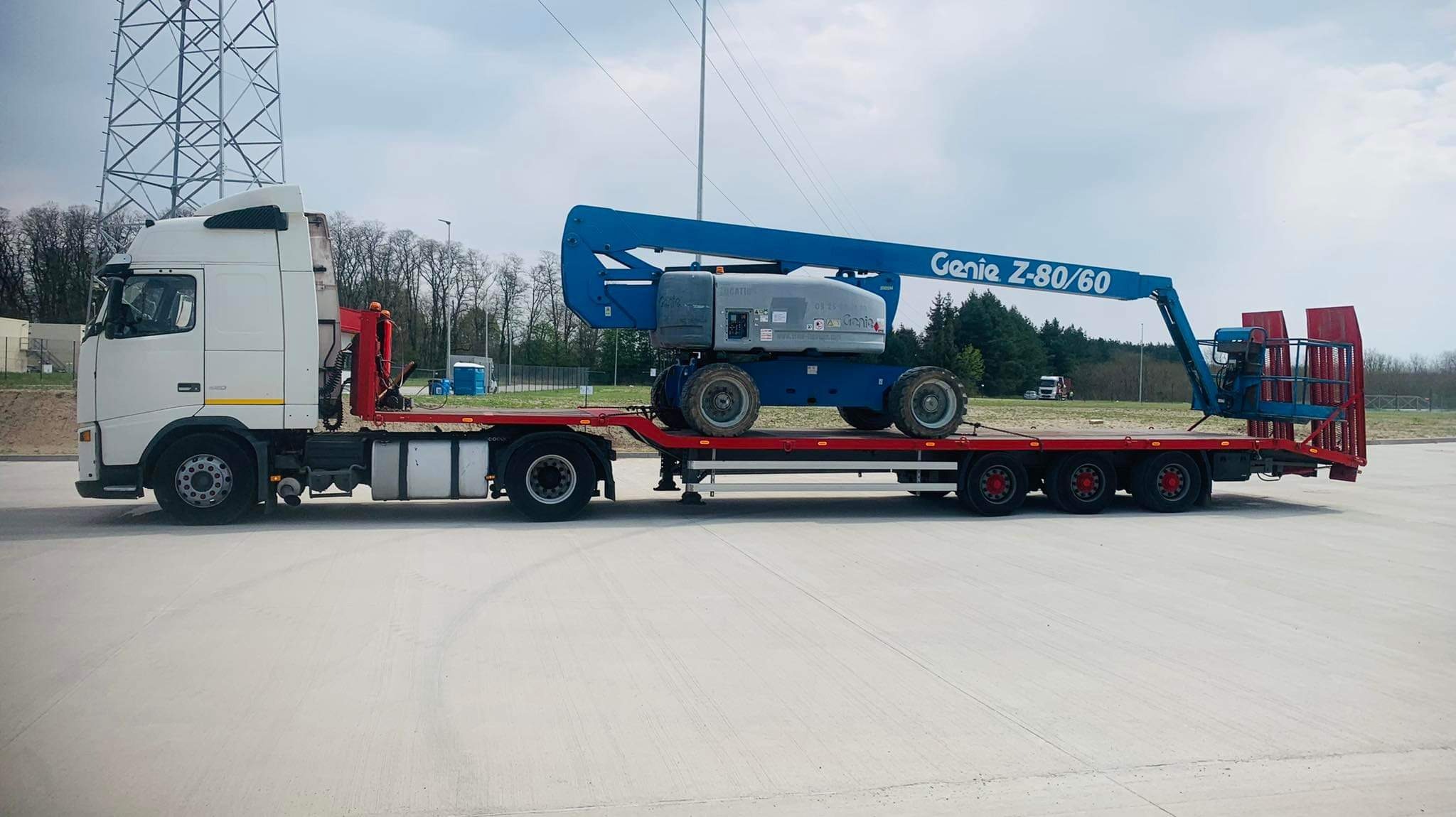 Biała ciężarówka z naczepą typu laweta przewozi niebieski podnośnik koszowy Genie Z-80/60 na tle słupa wysokiego napięcia i lasu.