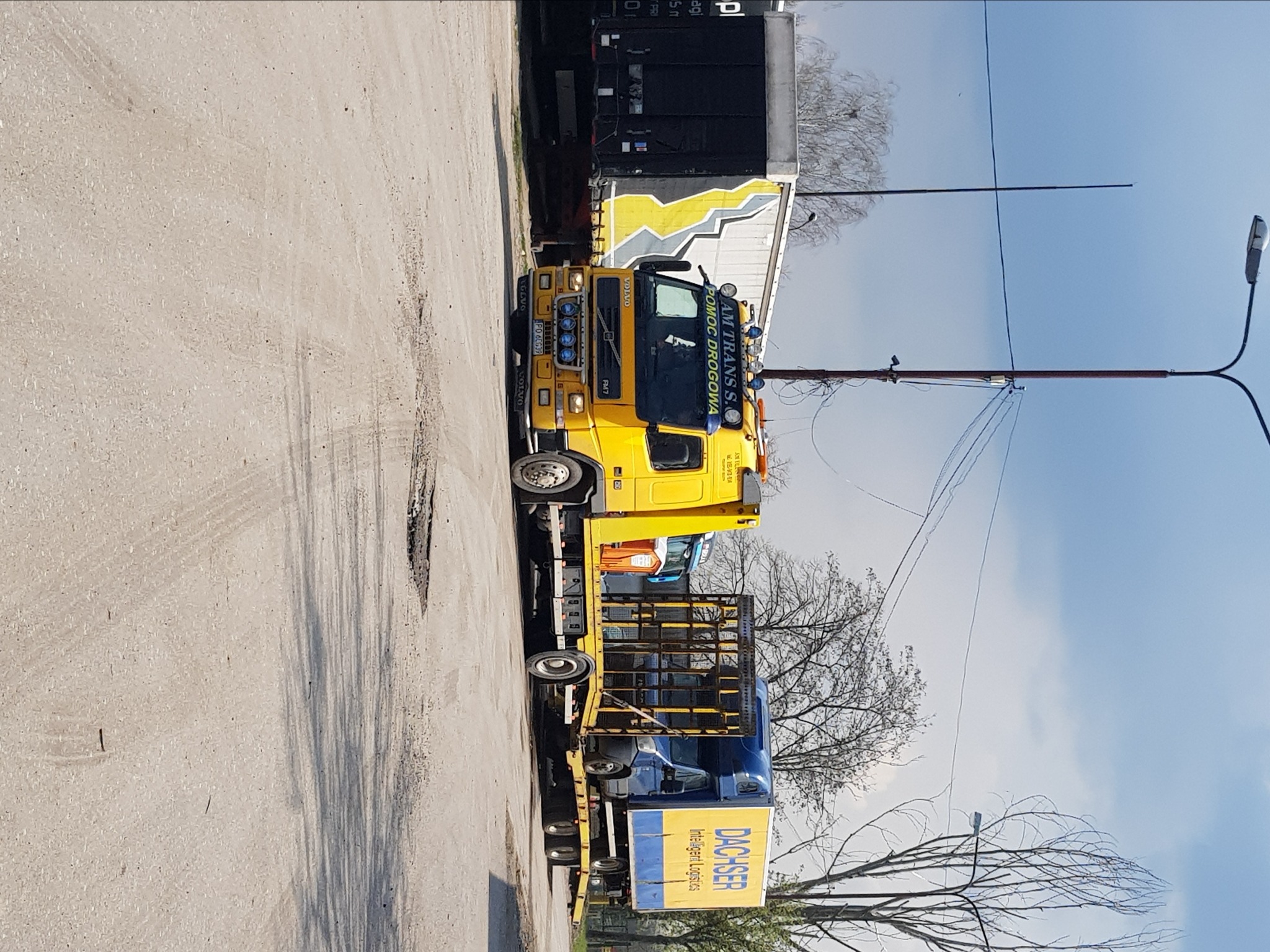 Żółta ciężarówka pomocy drogowej AM Trans S. z Poznania, z niebieskim napisem POMOC DROGOWA, przewożąca na lawecie niebieską ciężarówkę Dachser na szarym placu.