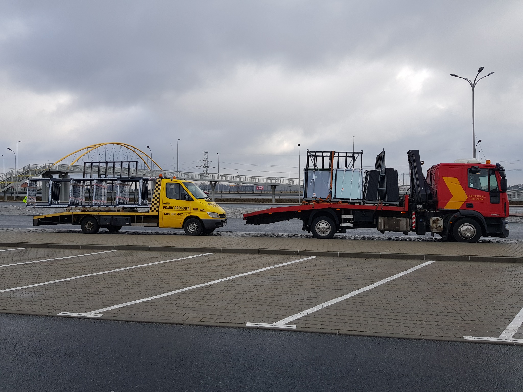 Dwa pojazdy pomocy drogowej, żółty i czerwony, z ładunkiem szyb na platformach, stoją na parkingu z widocznym żółtym mostem dla pieszych w tle, pochmurne niebo.