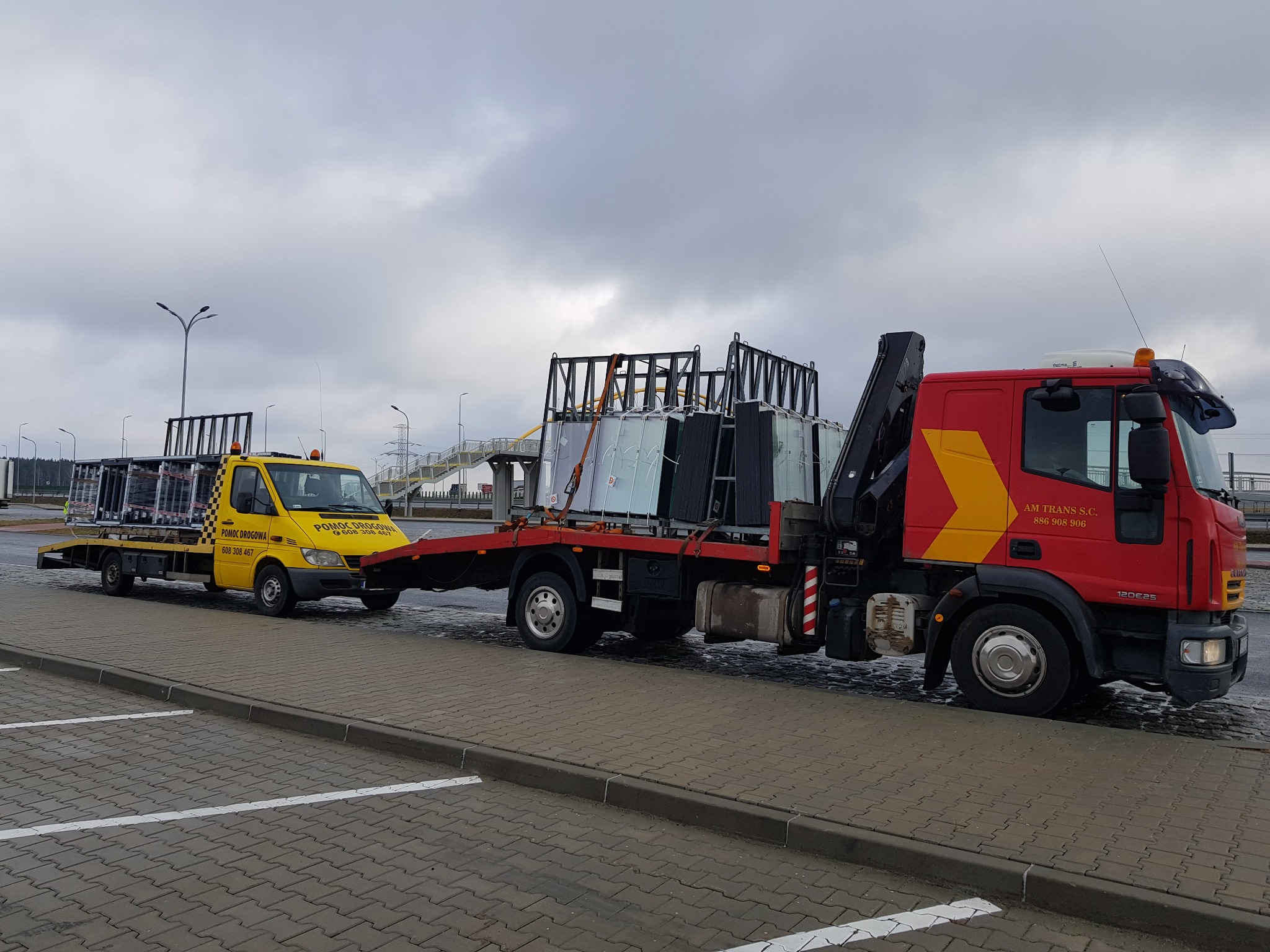 Żółta laweta holuje czerwoną ciężarówkę z dźwigiem, obie załadowane ramami z szybami na parkingu z kostki brukowej w pochmurny dzień.