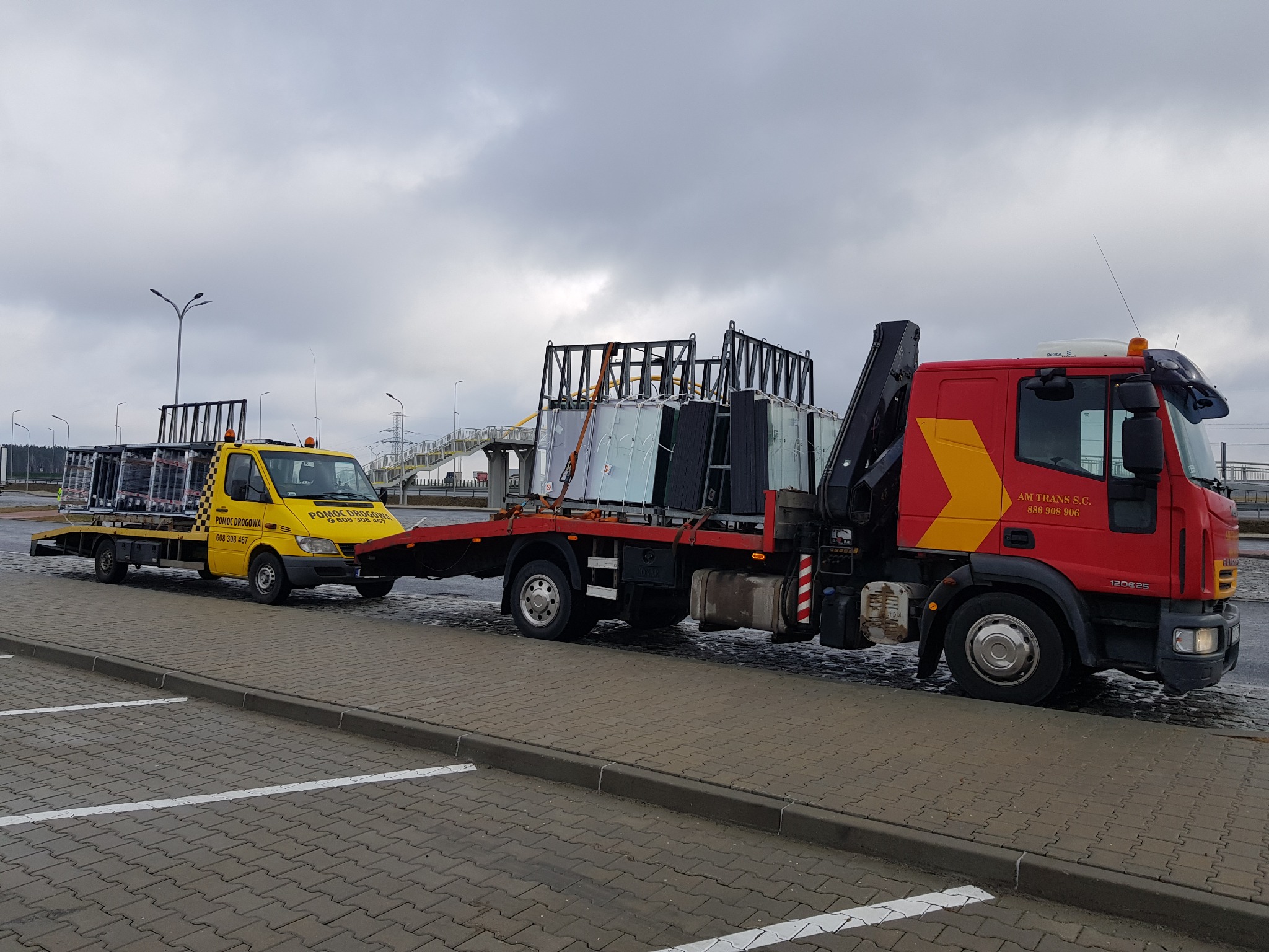 Żółta laweta holuje czerwoną lawetę z ładunkiem szklanych tafli na parkingu, pochmurne niebo w tle.