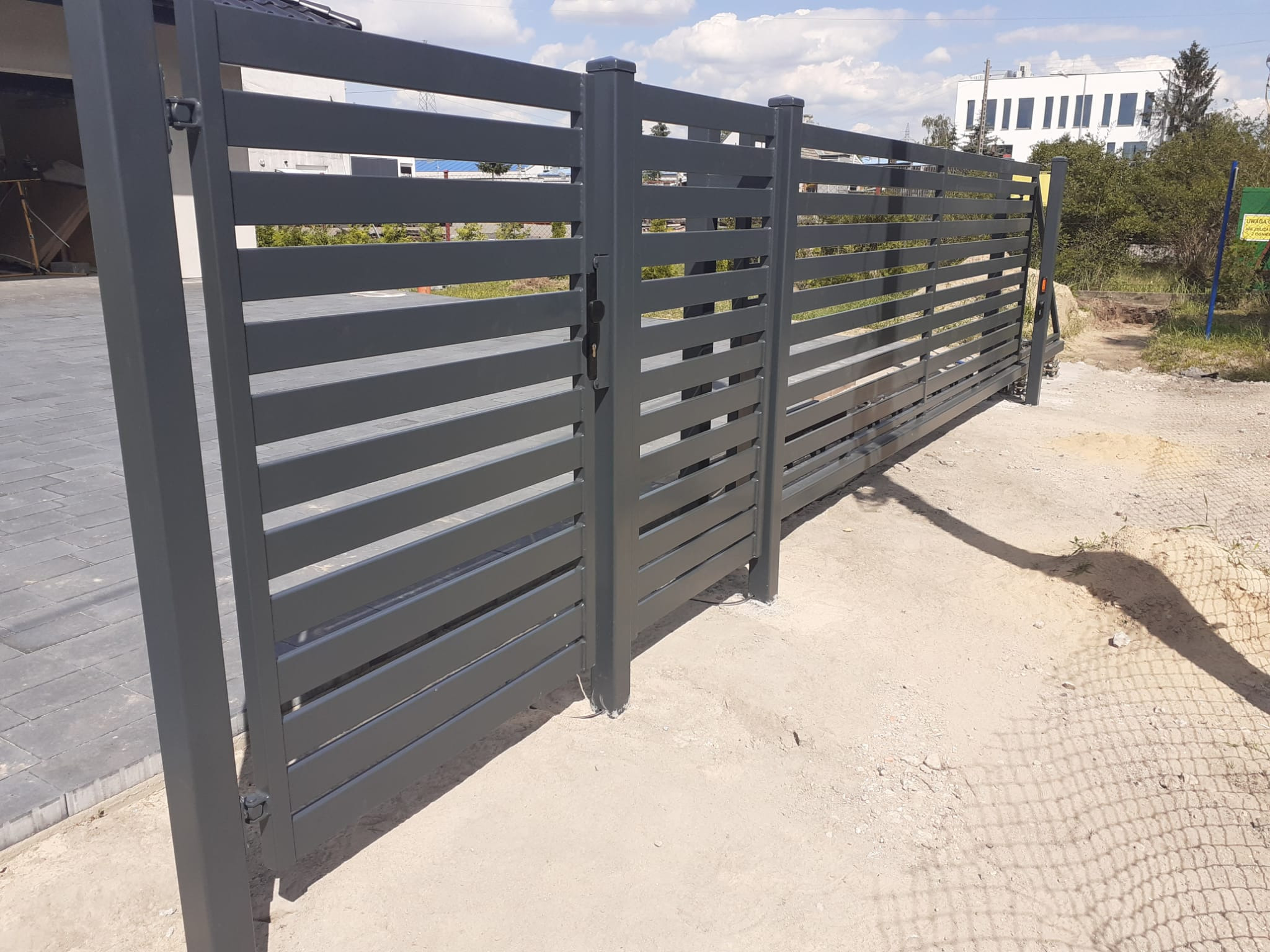 Nowoczesna, przesuwna brama wjazdowa z poziomych, szarych profili, widoczna z zewnątrz posesji na tle budynku i zieleni.