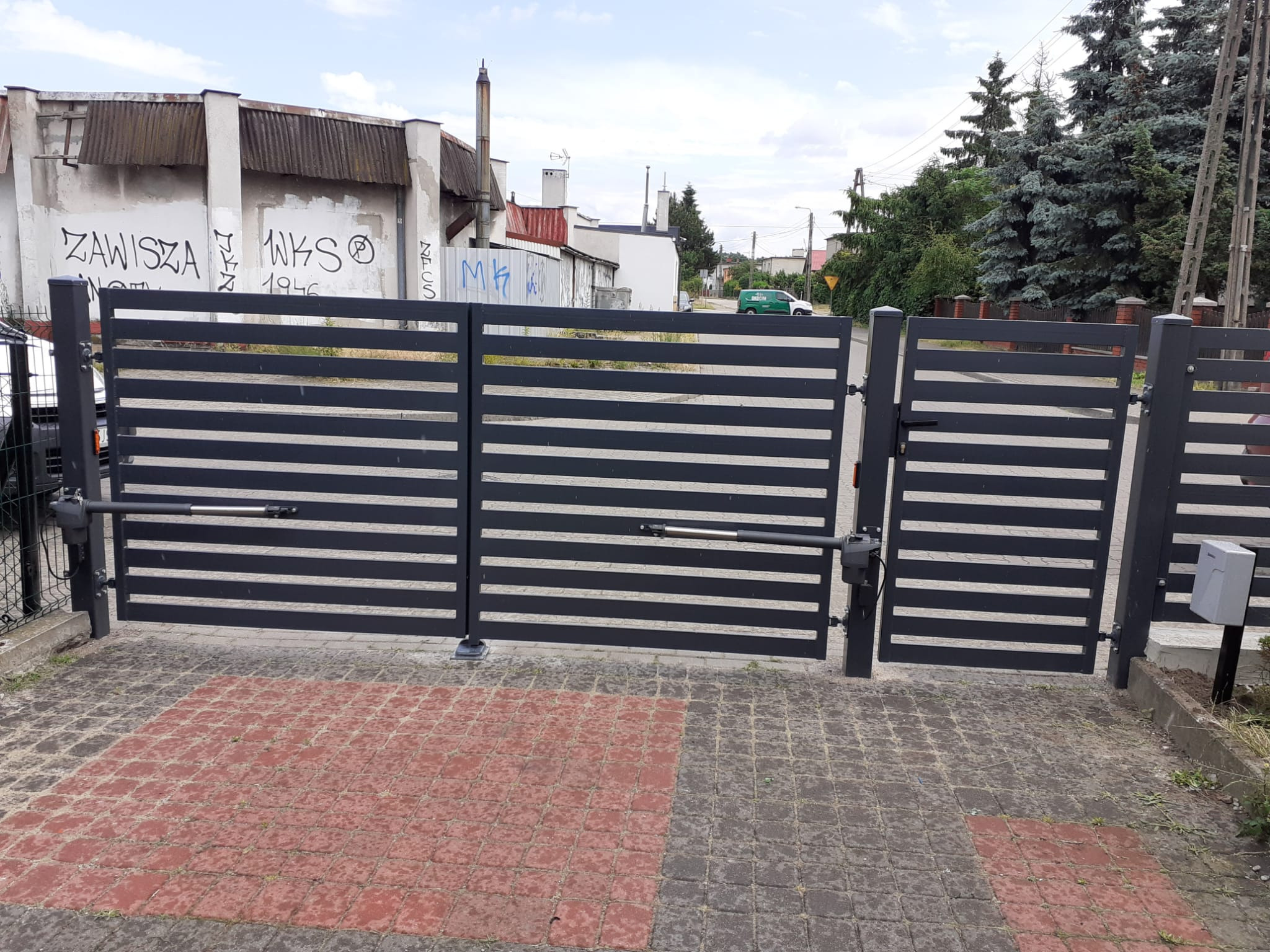 Nowoczesna, pozioma brama wjazdowa w kolorze grafitowym z automatyką, na posesji z kostką brukową w dwóch odcieniach, w tle zabudowania z graffiti.