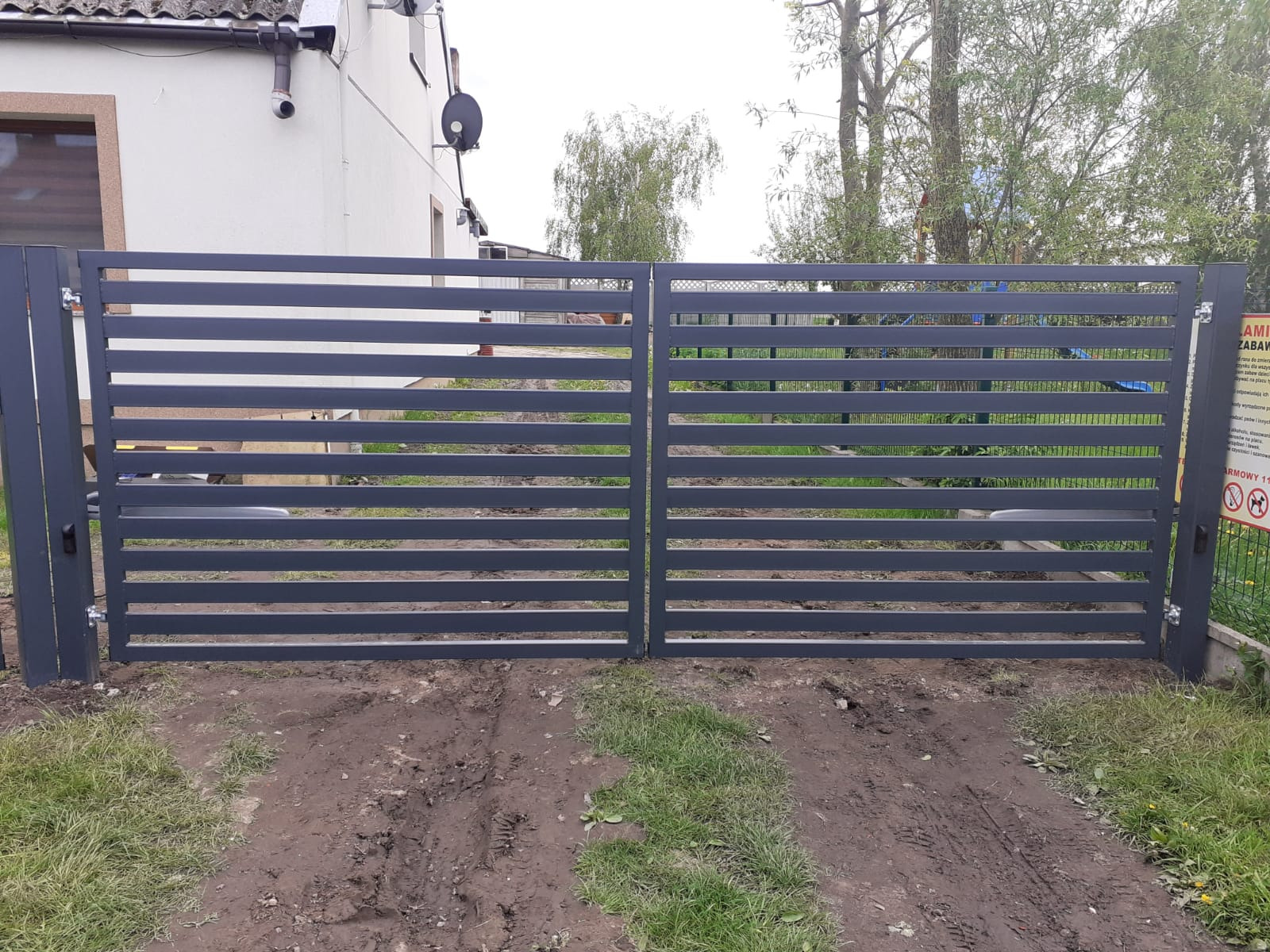 Nowoczesna, pozioma brama dwuskrzydłowa w kolorze grafitowym, z widocznym mechanizmem otwierania, osadzona na trawniku z błotnistymi śladami opon.
