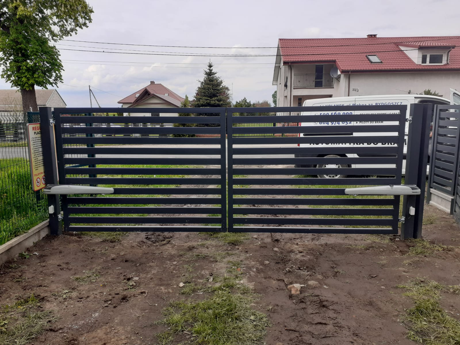 Automatyczna brama wjazdowa z poziomymi przęsłami w kolorze antracytowym, z dwoma siłownikami, na tle domu z czerwoną dachówką i firmowego samochodu dostawczego.