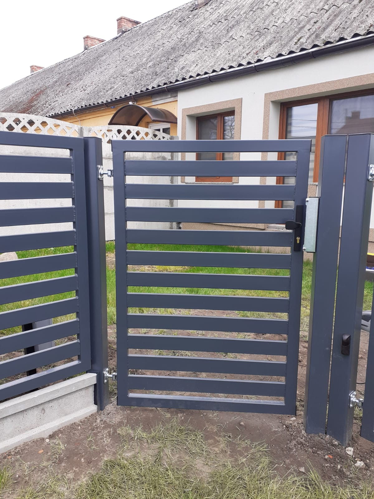 Nowoczesna, szara brama wjazdowa z poziomymi profilami, z widoczną klamką i zamkiem, osadzona na betonowej podmurówce, z domem w tle.