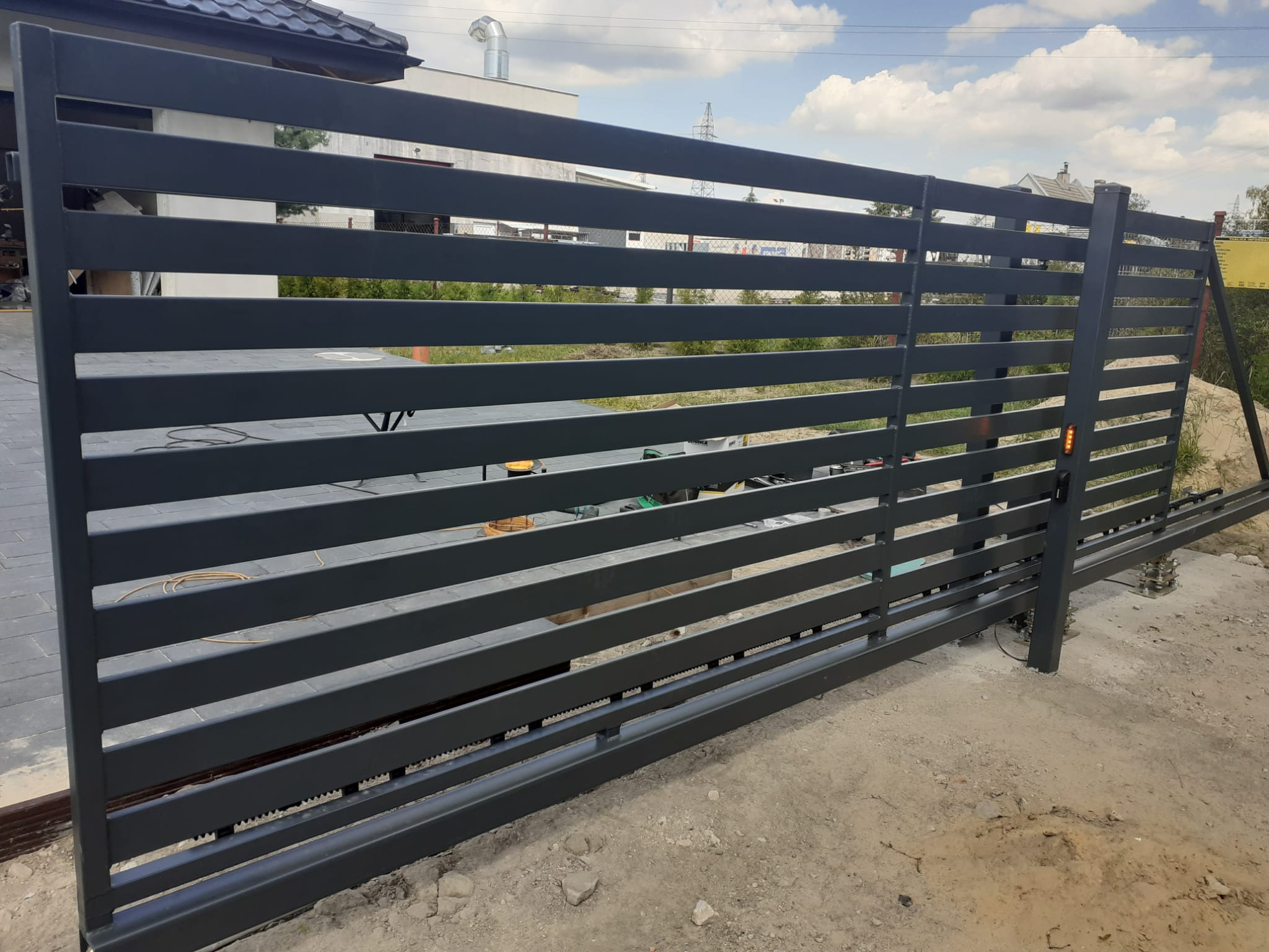 Nowoczesna, przesuwna brama wjazdowa z poziomych, grafitowych profili, widoczne elementy automatyki i napędu, teren wokół w trakcie prac wykończeniowych.