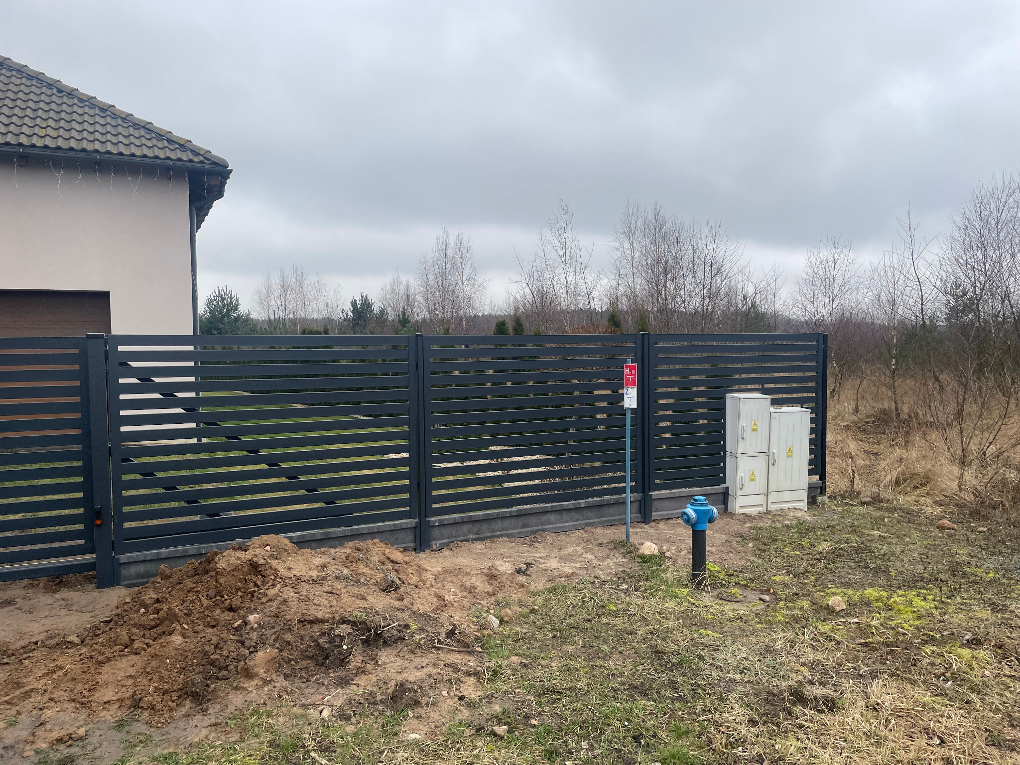 Nowoczesne, poziome, szare ogrodzenie panelowe na podmurówce, świeżo po montażu, widoczny kopiec ziemi, w tle dom z brązową dachówką i zarośla pod pochmurnym niebem.
