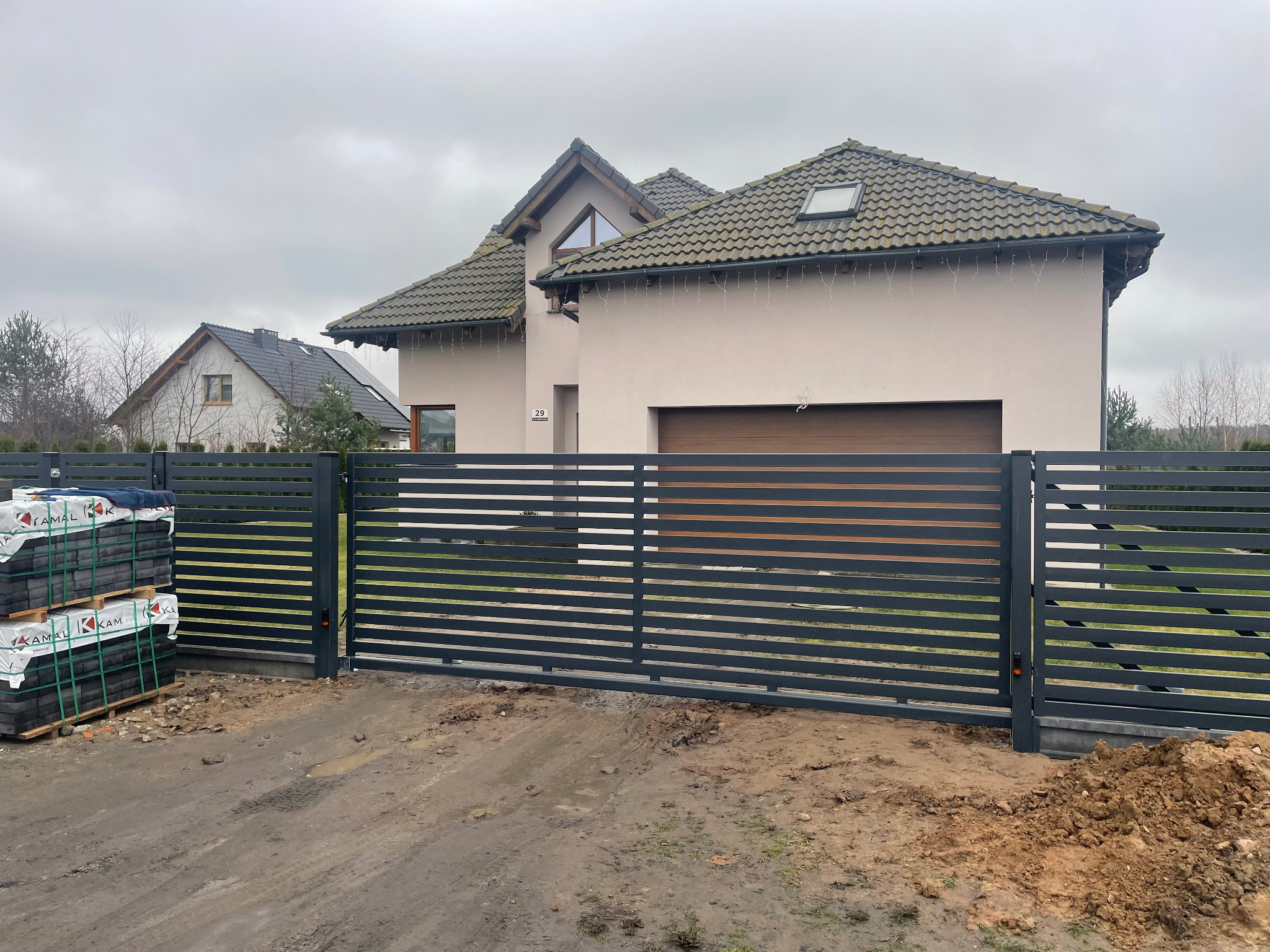 Nowoczesna, pozioma brama wjazdowa z szarych profili stalowych na tle domu z beżową elewacją i brązową bramą garażową, obok sterta kostki brukowej przed ułożeniem.