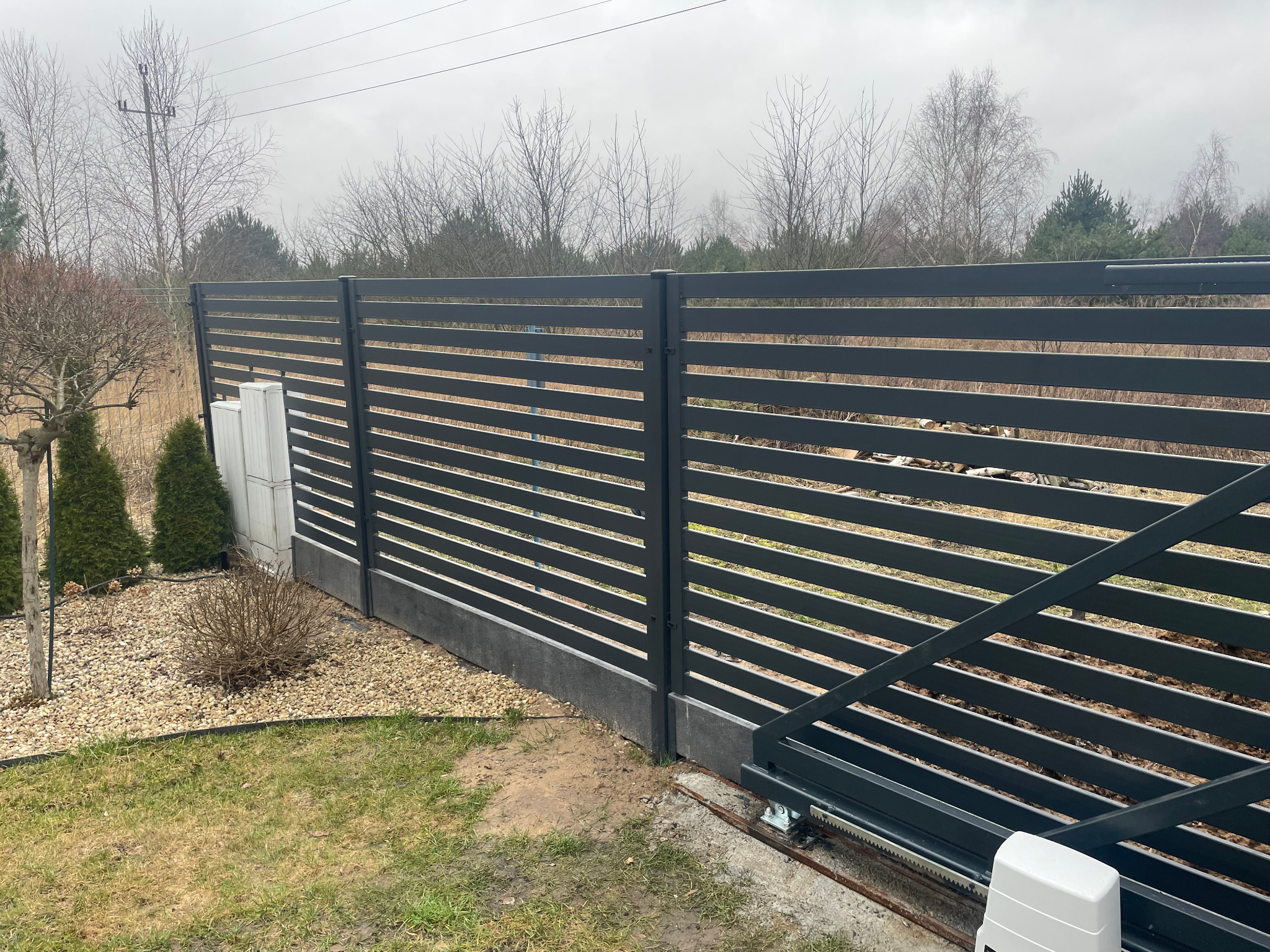 Nowoczesna, pozioma, szara brama przesuwna z automatyką, widok na ogród z kamieniami i roślinami, pochmurne niebo w tle.