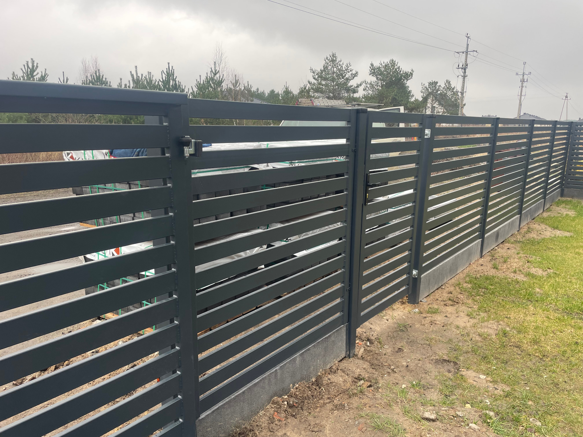 Nowoczesna, pozioma, grafitowa brama wjazdowa z metalowymi przęsłami i betonową podmurówką, widoczna z boku na tle pochmurnego nieba i drzew.