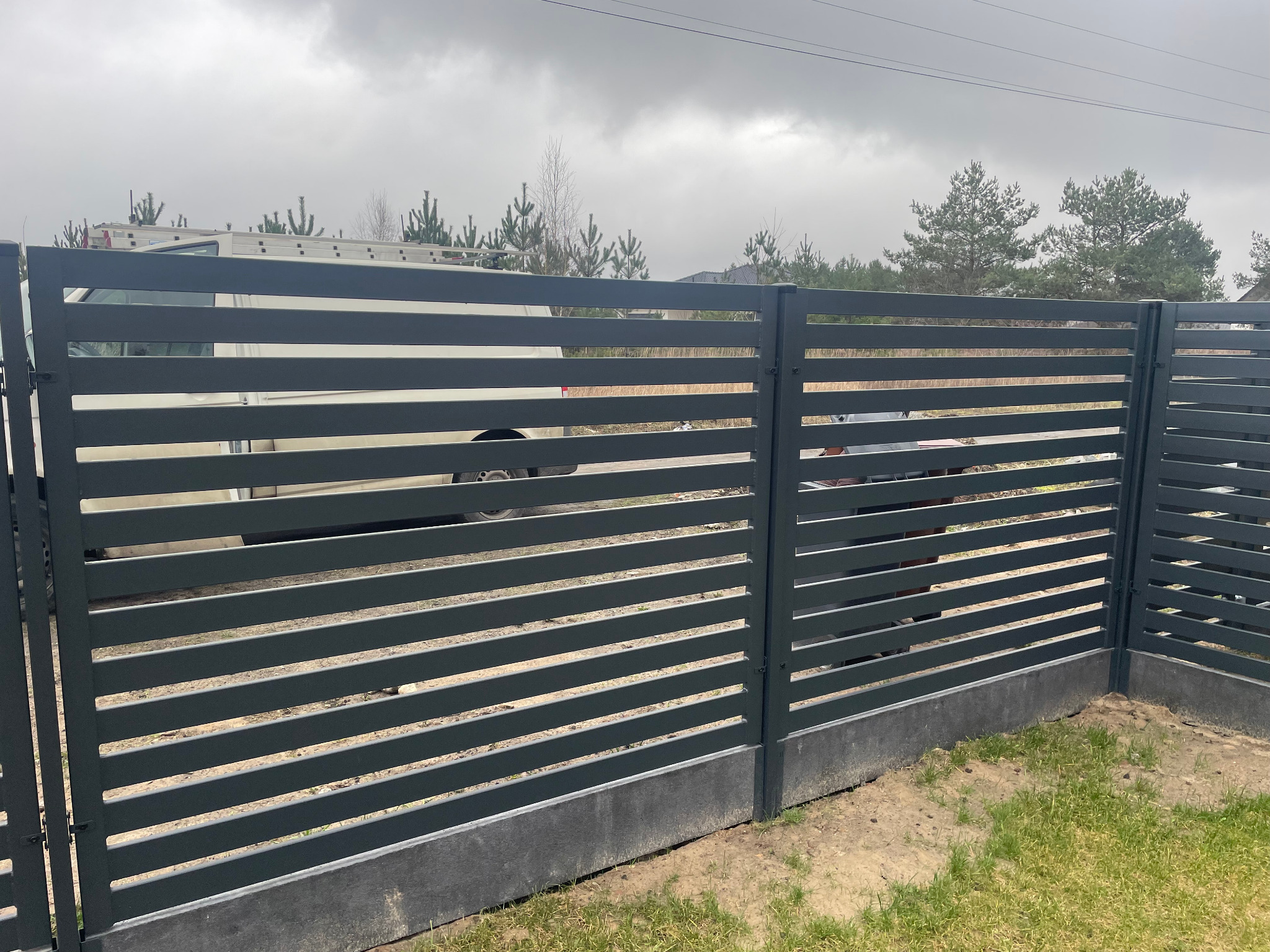 Nowoczesne, poziome ogrodzenie panelowe w kolorze grafitowym z podmurówką betonową, widok z zewnątrz posesji w pochmurny dzień. W tle zaparkowany biały bus z drabiną na dachu.