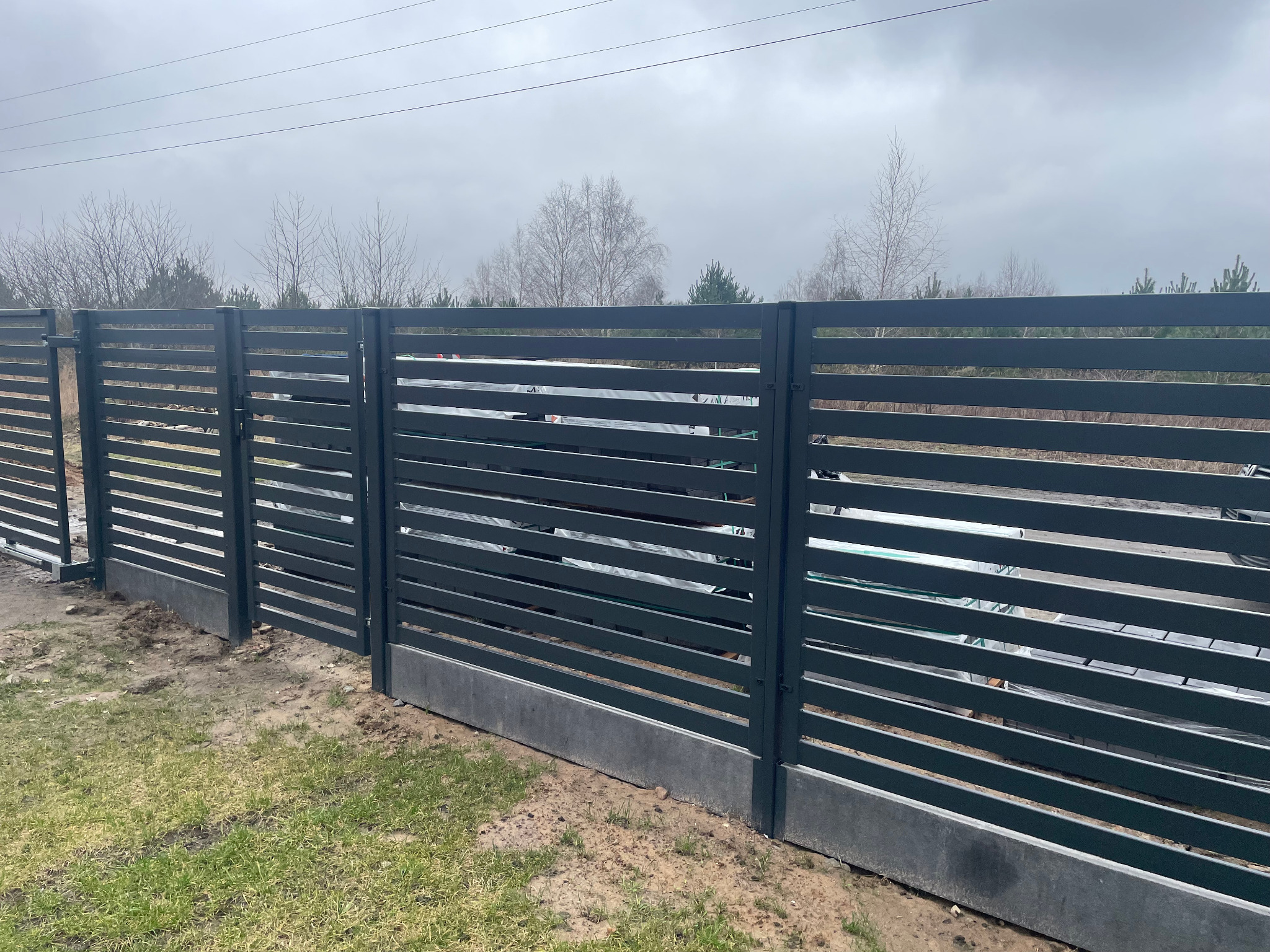Nowoczesne, poziome ogrodzenie panelowe w kolorze grafitowym z betonową podmurówką na trawiastym terenie, widok z zewnątrz.