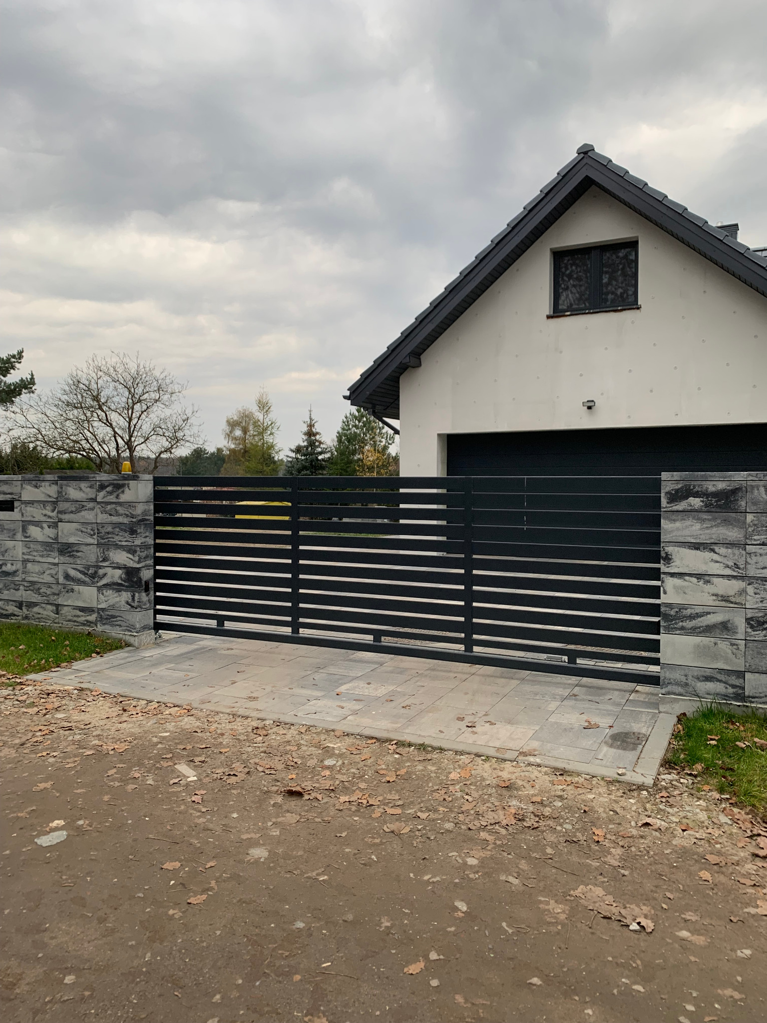 Nowoczesna, przesuwna brama wjazdowa z poziomych profili, zamontowana na posesji z domem o białej elewacji i szarej kostce brukowej. Słupki bramy wykończone dekoracyjnym kamieniem elewacyjnym.