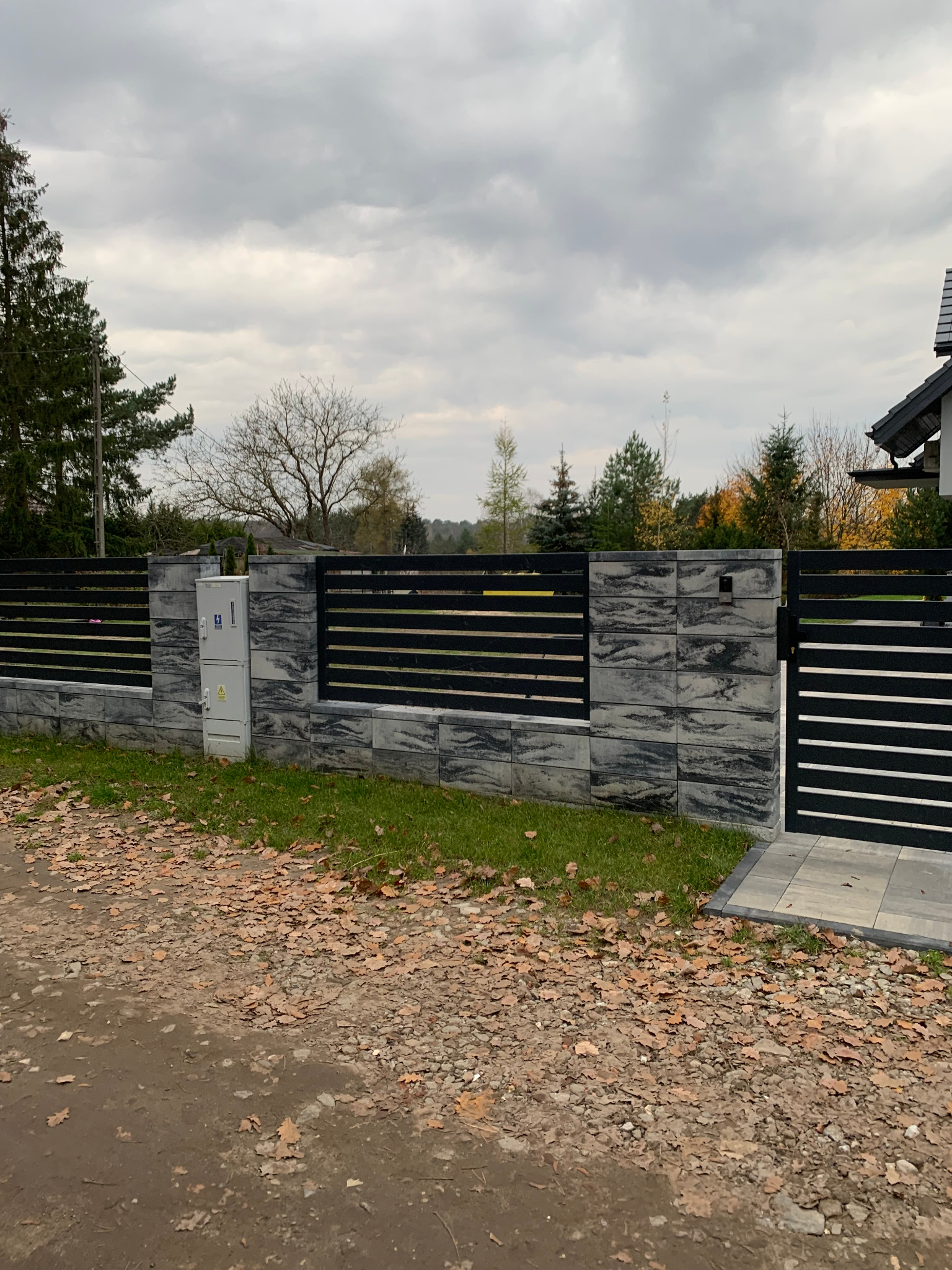 Nowoczesne ogrodzenie panelowe z czarnymi, poziomymi przęsłami i szarymi słupkami z betonu architektonicznego, brama wjazdowa z identycznym wypełnieniem, skrzynka elektryczna na słupku, jesienne...