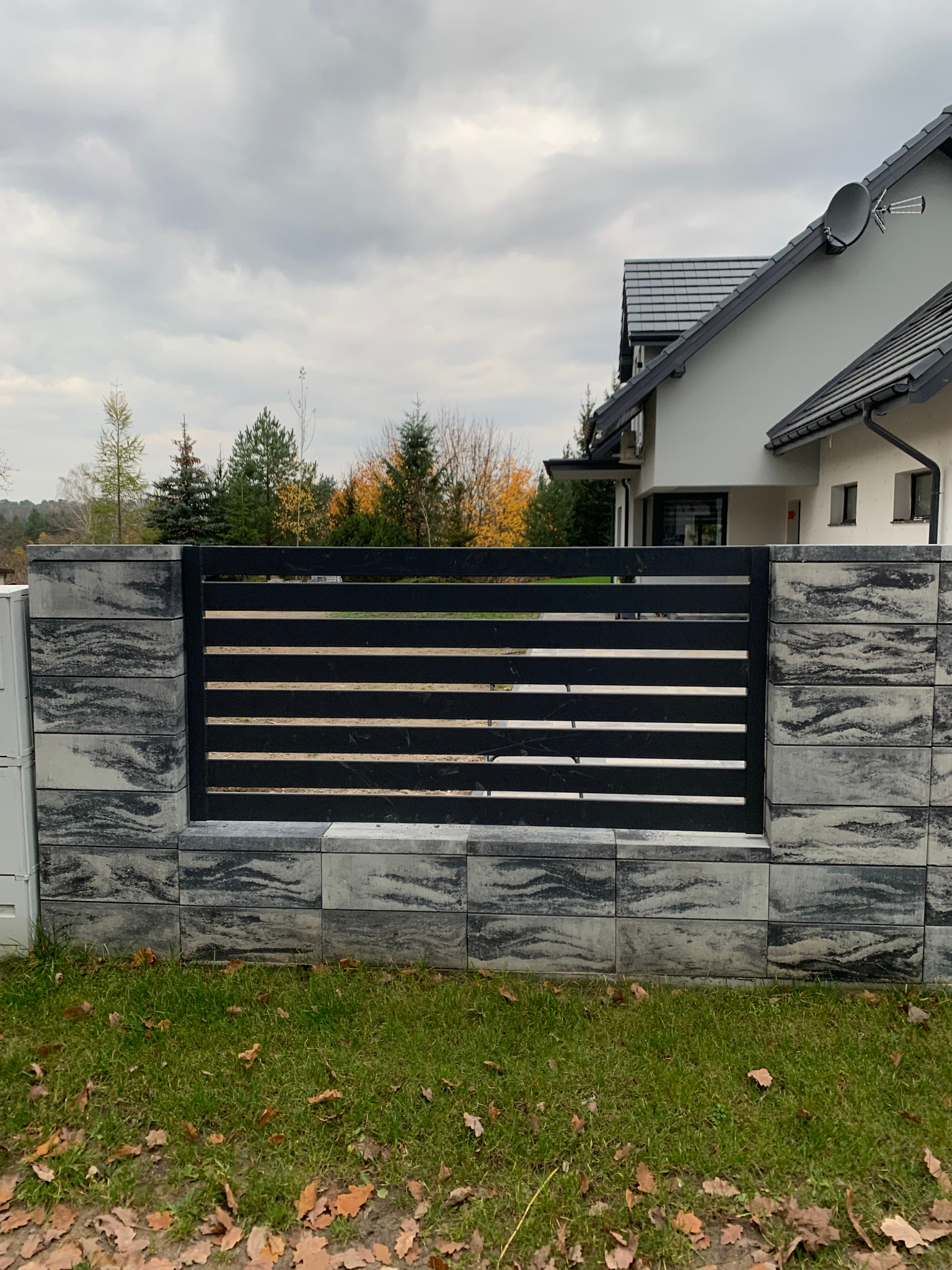 Nowoczesne ogrodzenie panelowe z elementami betonowymi o marmurkowym wzorze, widok na dom z anteną satelitarną w tle, jesienne liście na trawie.