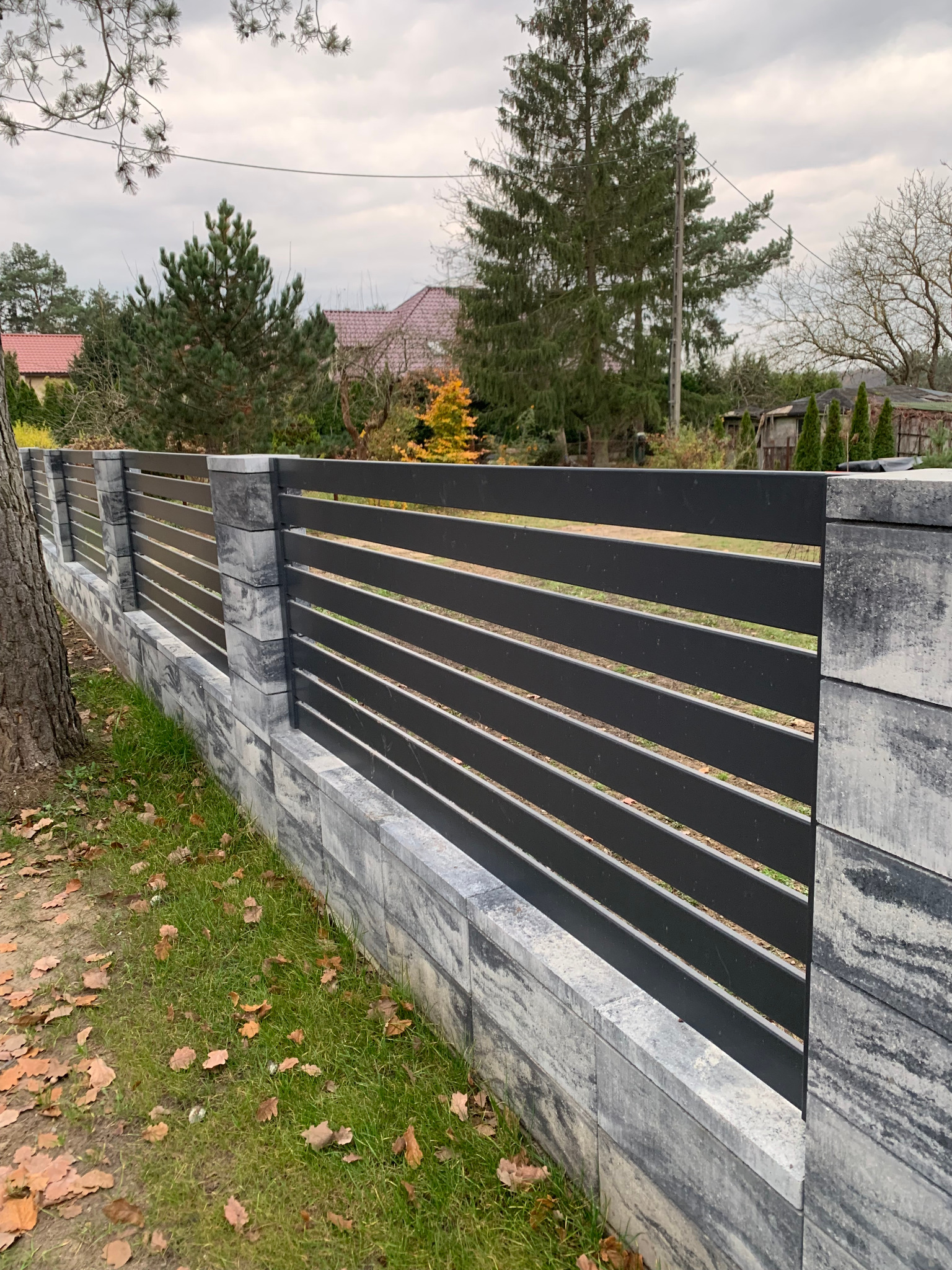 Nowoczesne ogrodzenie z poziomych, grafitowych paneli zamontowanych na murku z szarej kostki imitującej kamień, widok z poziomu gruntu.