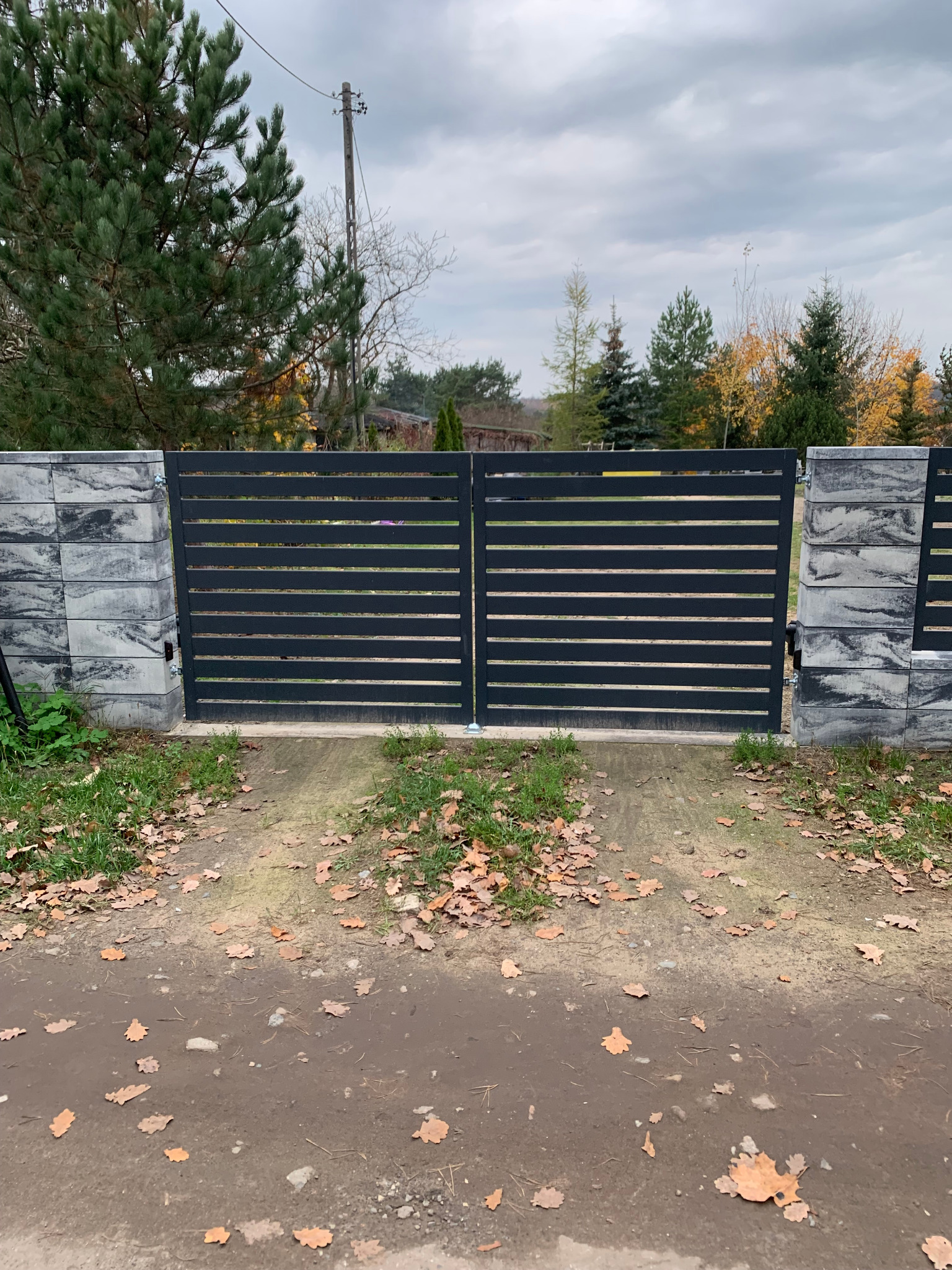 Nowoczesna, pozioma brama wjazdowa z szarego metalu, osadzona na betonowych słupkach z fakturą kamienia, na tle jesiennego krajobrazu z opadłymi liśćmi.