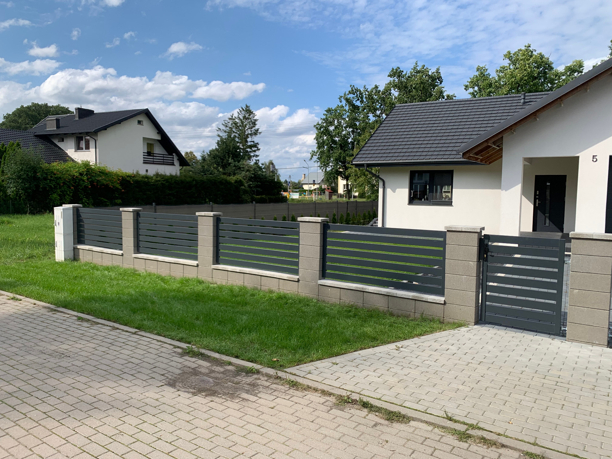 Nowoczesne ogrodzenie panelowe w kolorze grafitowym z bramką, osadzone na betonowej podmurówce i słupkach, na tle domów jednorodzinnych i błękitnego nieba z chmurami.