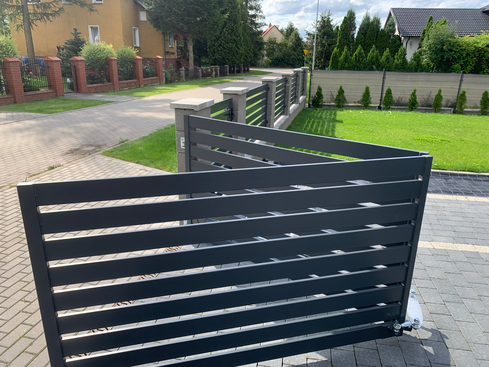 Nowoczesna, grafitowa brama przesuwna z poziomymi panelami, częściowo otwarta, widoczny mechanizm jezdny, na tle kostki brukowej i ogrodzenia panelowego.