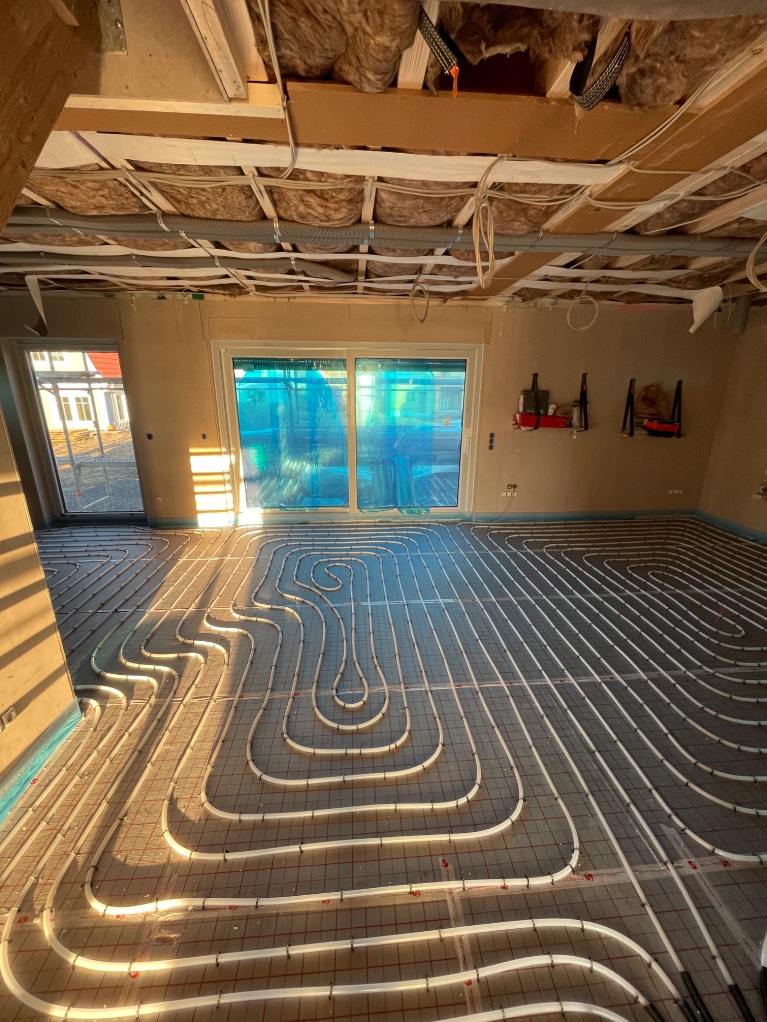 Wnętrze w trakcie instalacji ogrzewania podłogowego, widoczne białe rury ułożone w pętle na specjalnej macie, promienie zachodzącego słońca wpadają przez okno balkonowe.