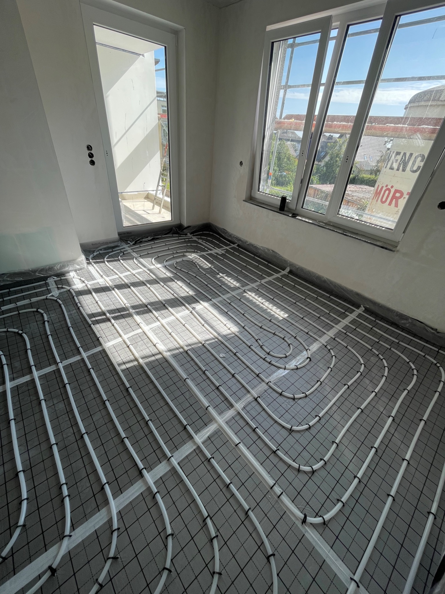 Białe rury ogrzewania podłogowego ułożone na czarnej siatce w surowym pomieszczeniu z oknem i wyjściem na balkon.