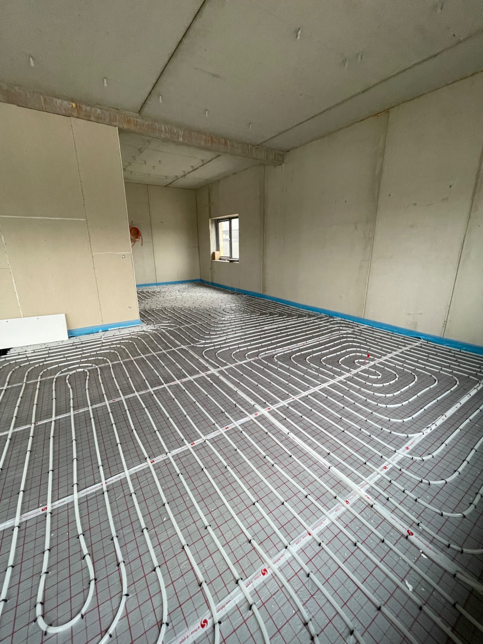 Rozłożone rury ogrzewania podłogowego na folii z siatką w surowym wnętrzu, widoczne łączenia rur i fragment okna.