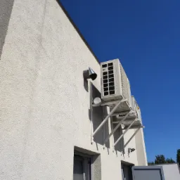 Dwa klimatyzatory zamontowane na zewnątrz budynku z białą elewacją, widoczne wsporniki montażowe, rura wentylacyjna i fragment okna, na tle błękitnego nieba.