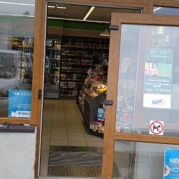 SERWIS JACEK MARTA PIECZONKA - Drewniane drzwi wejściowe do sklepu spożywczego częściowo zdemontowane, w tle widoczne półki z piwem i innymi produktami, na podłodze leży wkrętarka.