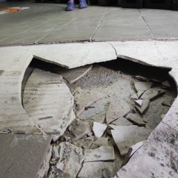 SERWIS JACEK MARTA PIECZONKA - Uszkodzona podłoga z płytek, widoczne fragmenty gruzu i podłoża, w tle stopy w kolorowych klapkach.