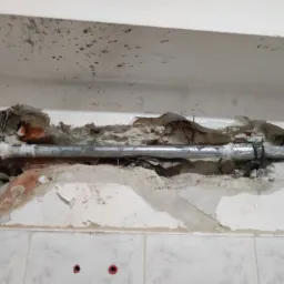 Rura kanalizacyjna w ścianie z uszkodzonym tynkiem i widoczną cegłą, fragment ściany wyłożony białymi płytkami.