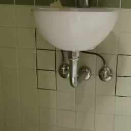 Biała umywalka zamontowana na kafelkowej ścianie, widoczne chromowane rury i bateria, fragment toalety w prawym dolnym rogu.