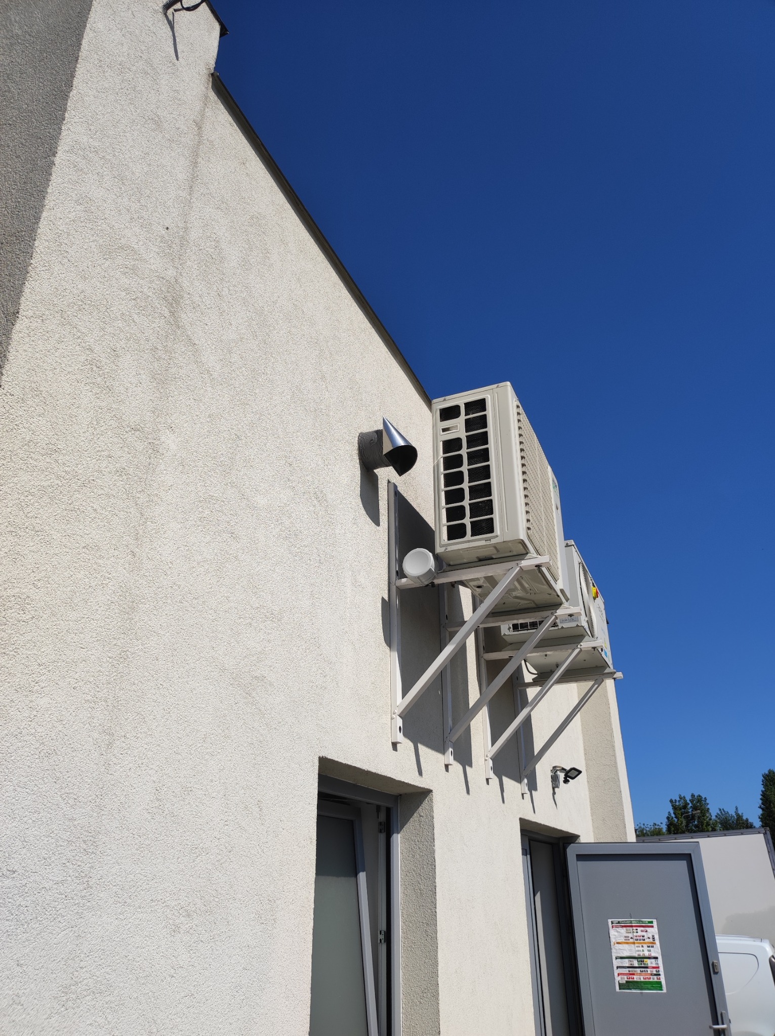 Dwa klimatyzatory zamontowane na zewnątrz budynku z białą elewacją, widoczne wsporniki montażowe, rura wentylacyjna i fragment okna, na tle błękitnego nieba.