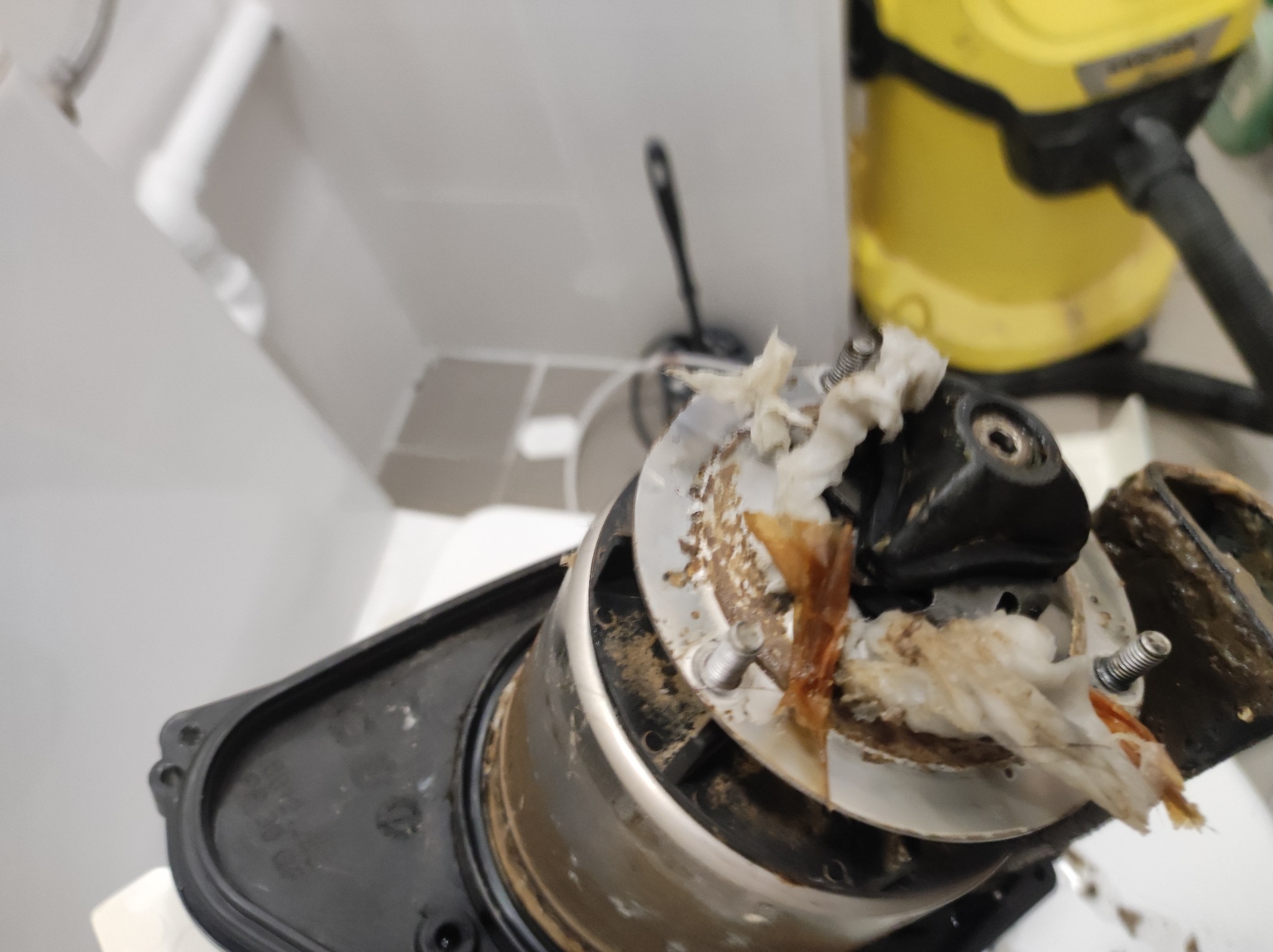 Zablokowany odpływ z fragmentami zanieczyszczeń, widoczne elementy mechanizmu odpływowego i żółty odkurzacz w tle.