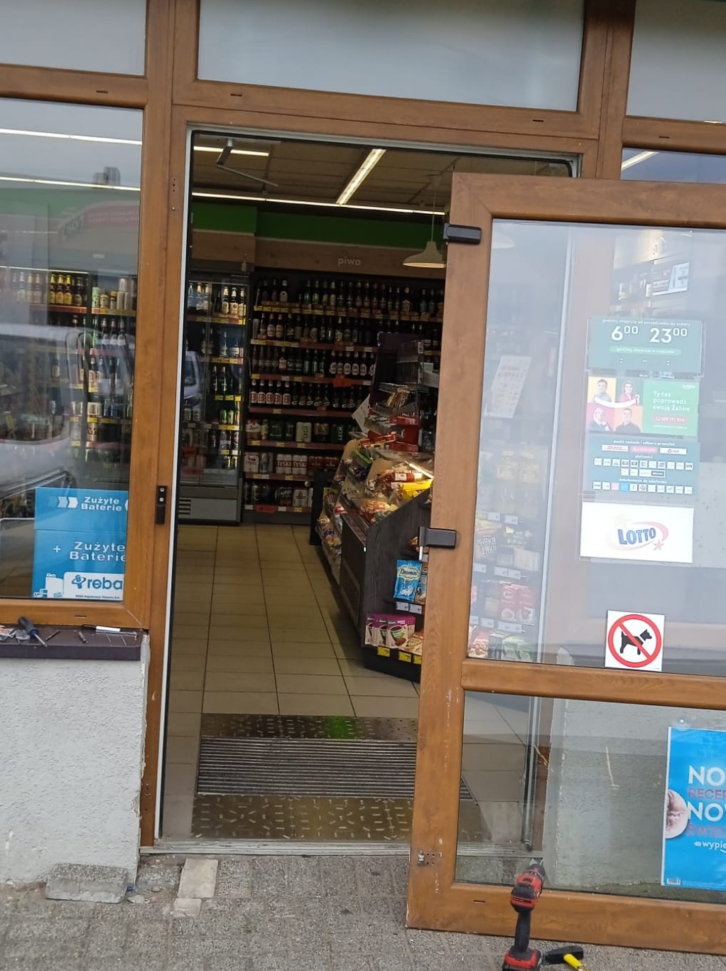 Drewniane drzwi wejściowe do sklepu spożywczego częściowo zdemontowane, w tle widoczne półki z piwem i innymi produktami, na podłodze leży wkrętarka.