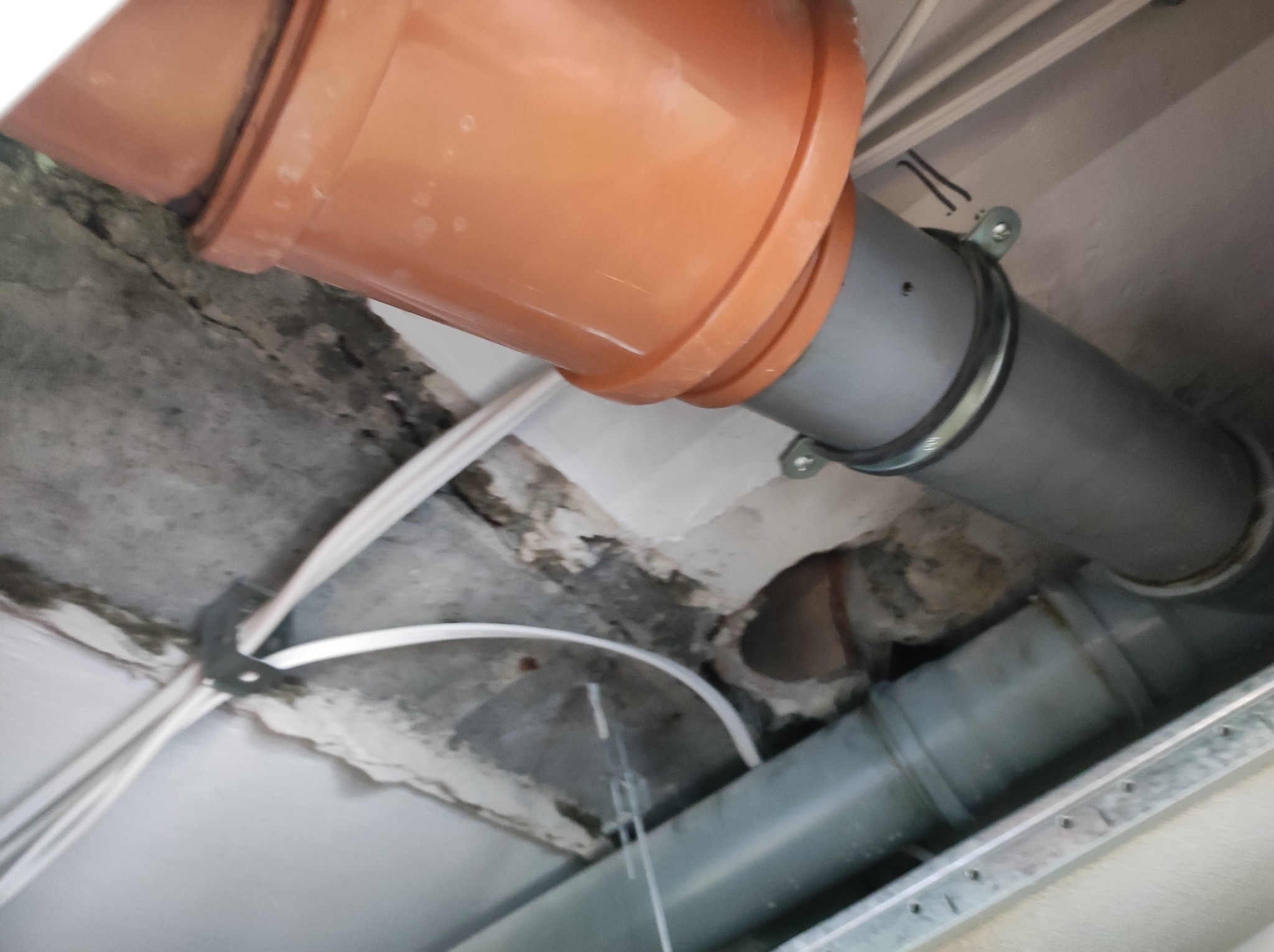 Pomarańczowa i szara rura kanalizacyjna przymocowana do sufitu za pomocą metalowych obejm, widoczne przewody elektryczne i fragmenty uszkodzonego sufitu.