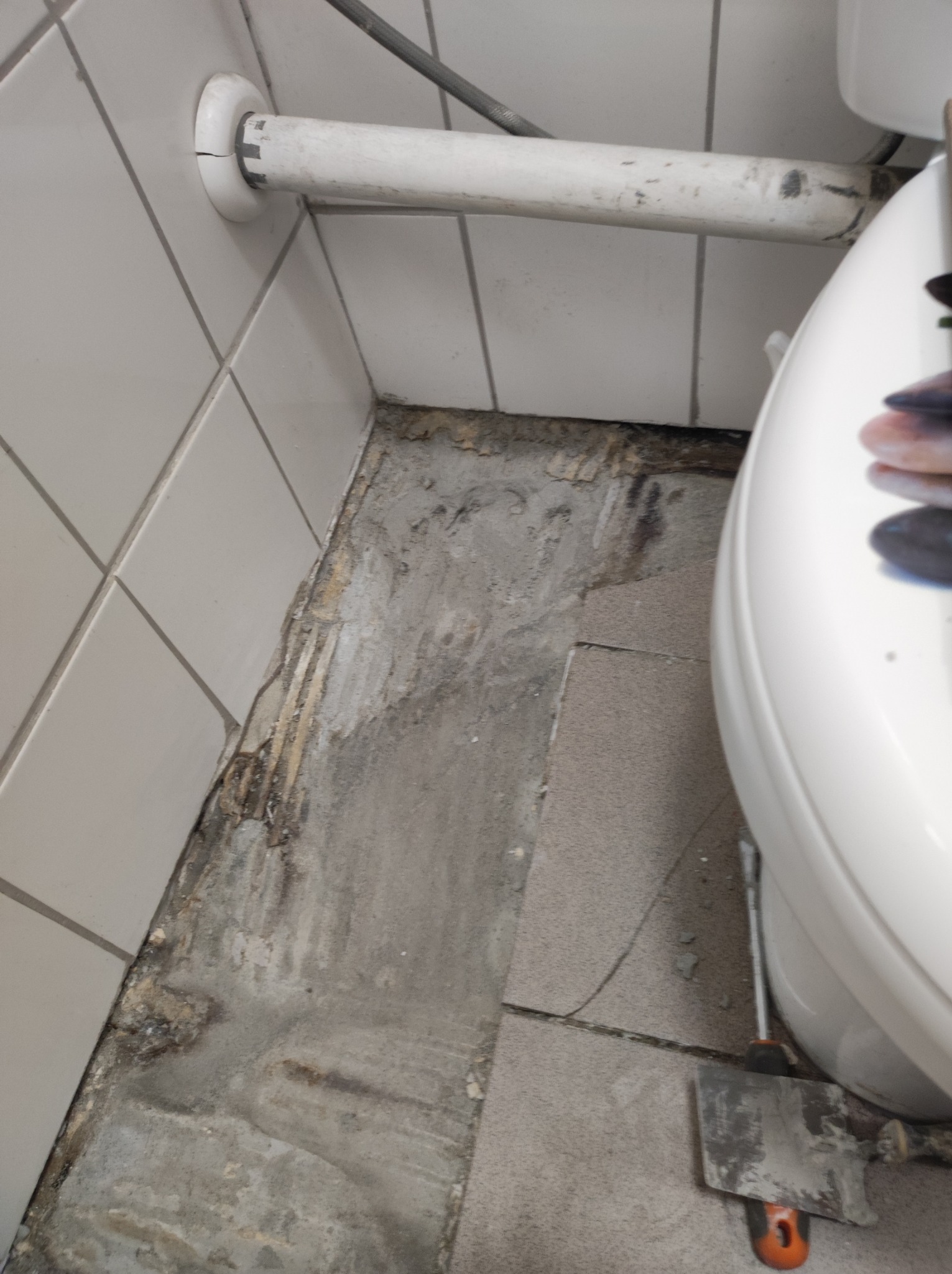 Odkryta podłoga w łazience w trakcie remontu, widoczne uszkodzenia i pozostałości po starej podłodze, biała rura przy ścianie i narzędzie do usuwania płytek obok muszli klozetowej.