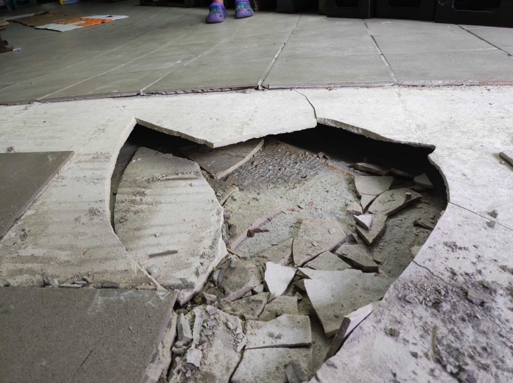 Uszkodzona podłoga z płytek, widoczne fragmenty gruzu i podłoża, w tle stopy w kolorowych klapkach.