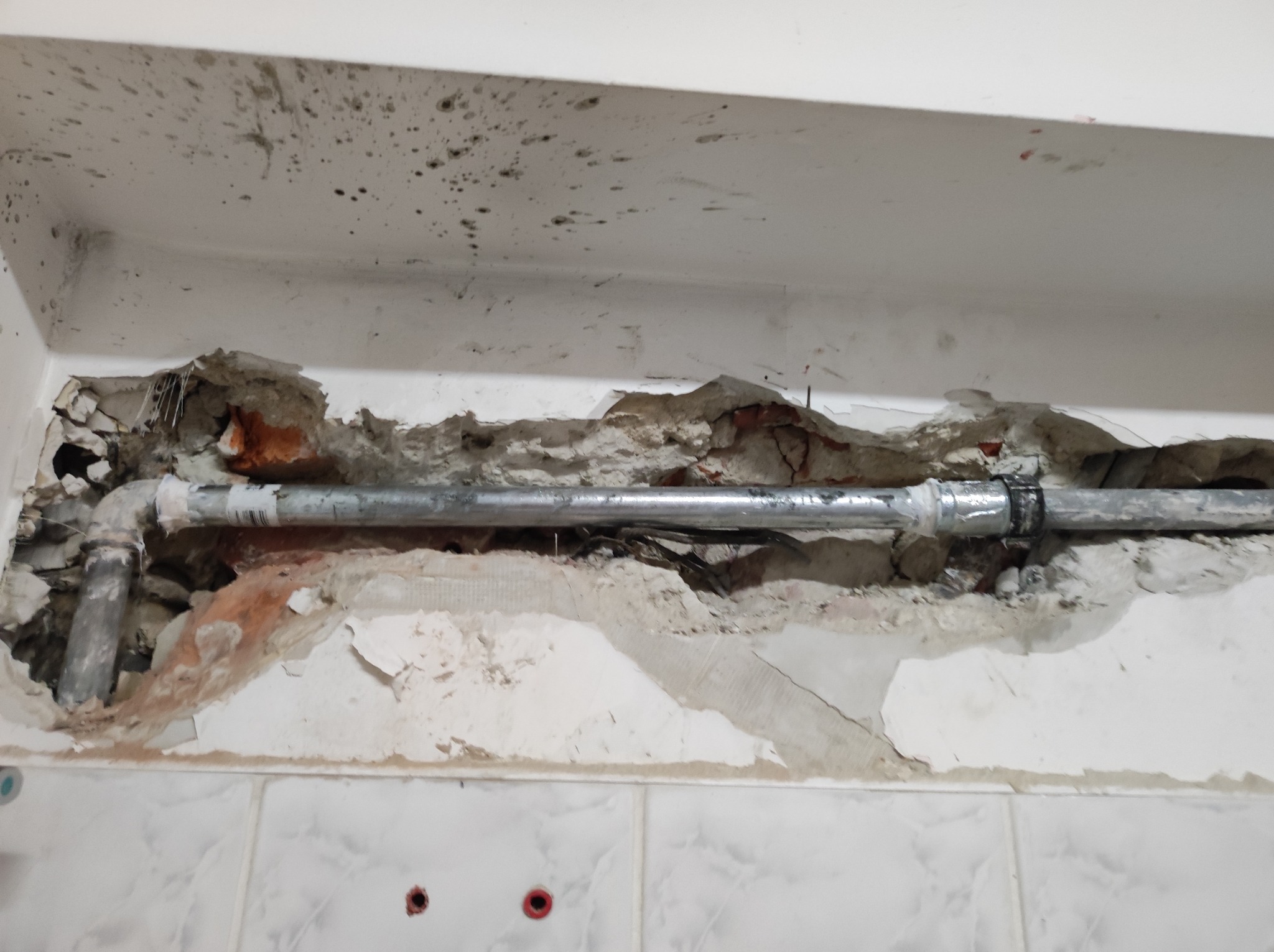 Rura kanalizacyjna w ścianie z uszkodzonym tynkiem i widoczną cegłą, fragment ściany wyłożony białymi płytkami.