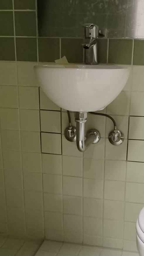 Biała umywalka zamontowana na kafelkowej ścianie, widoczne chromowane rury i bateria, fragment toalety w prawym dolnym rogu.